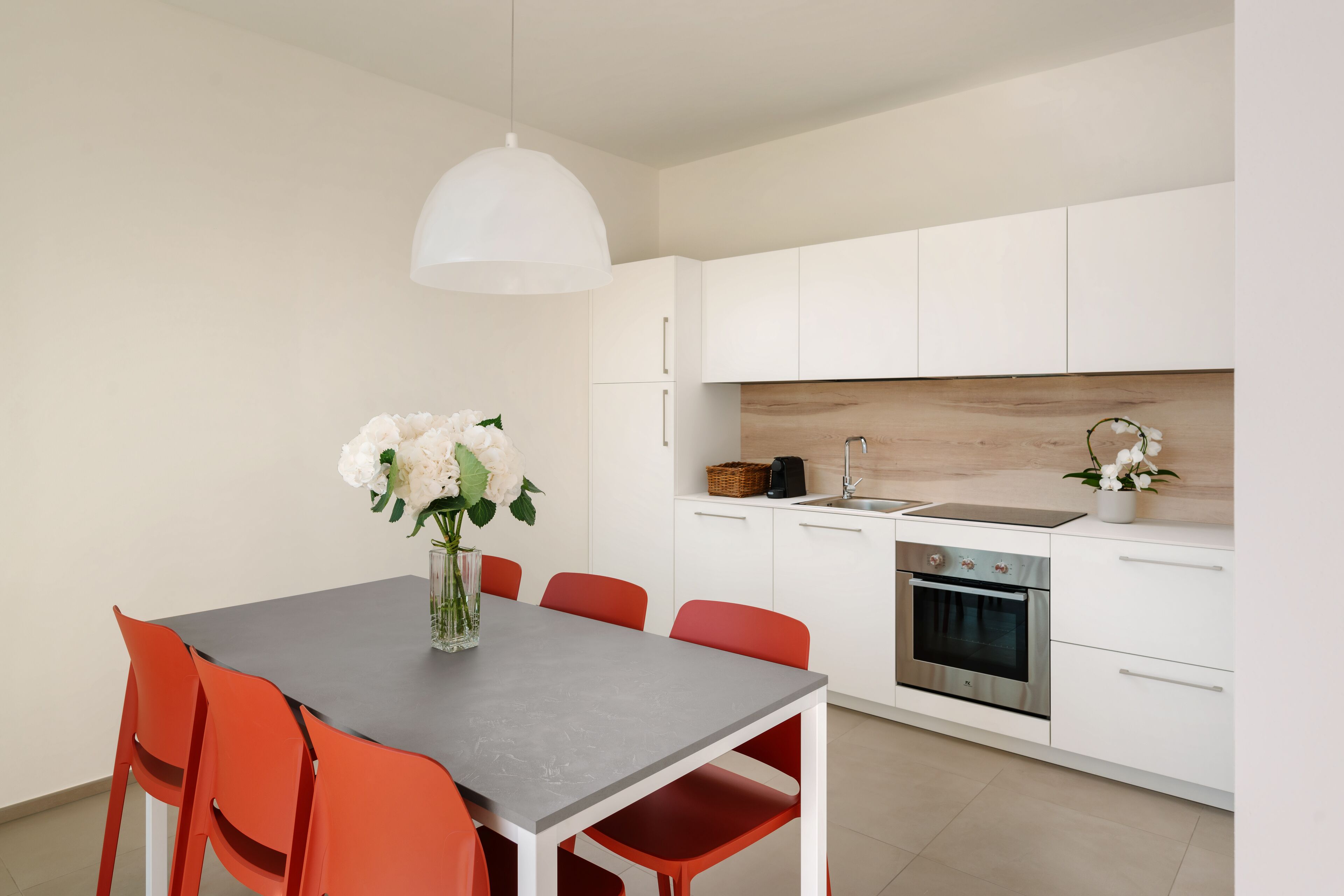 Eine moderne Küche mit weißen Schränken, einer grauen Arbeitsplatte und einem Edelstahlbackofen. Ein Esstisch mit roten Stühlen und eine Vase mit weißen Blumen sind ebenfalls vorhanden.