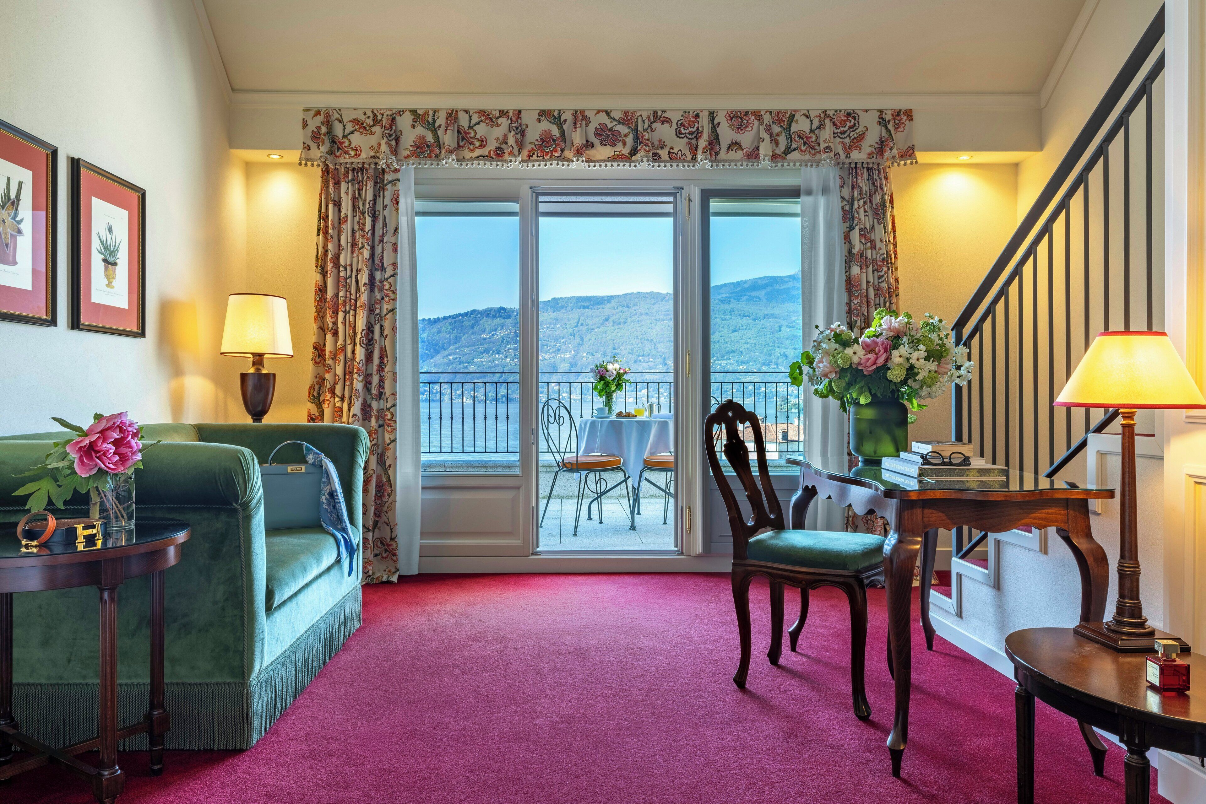 A room with a green sofa, a wooden chair, a table with a lamp, a balcony with a table and chairs, and a view of the hills.