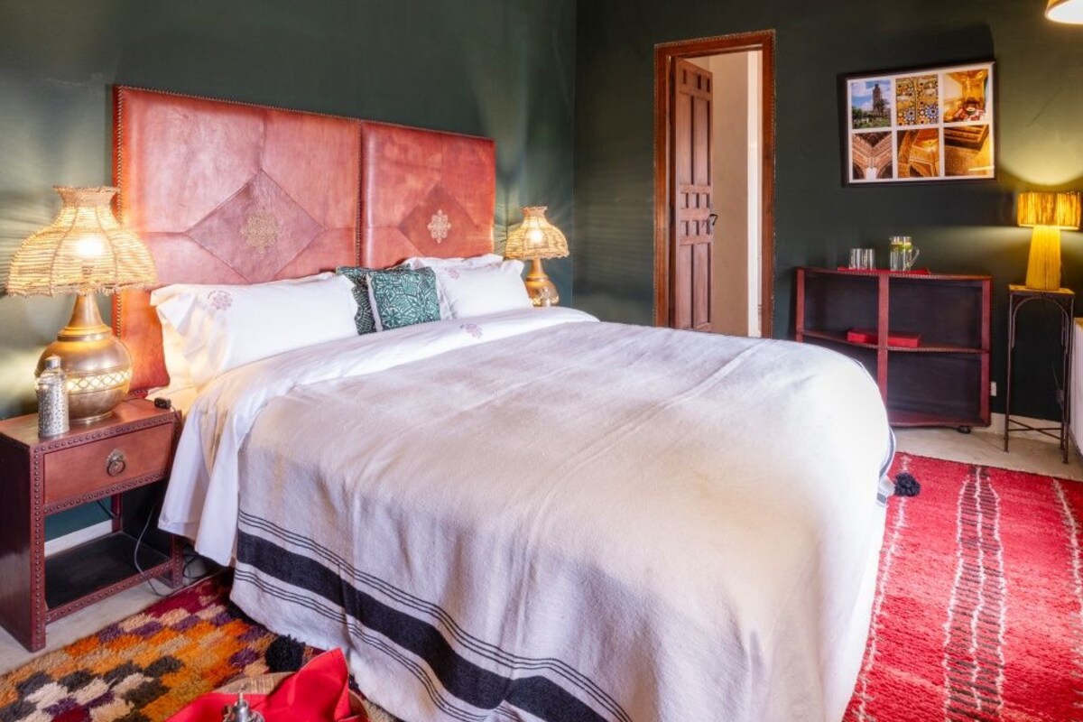 A bedroom with a large bed, two bedside tables with lamps, a wooden headboard, a red patterned rug, and a small shelf with items.