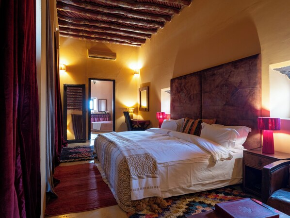 Habitación de hotel con una cama grande, techo de madera, alfombra roja y una pequeña mesa con una lámpara.