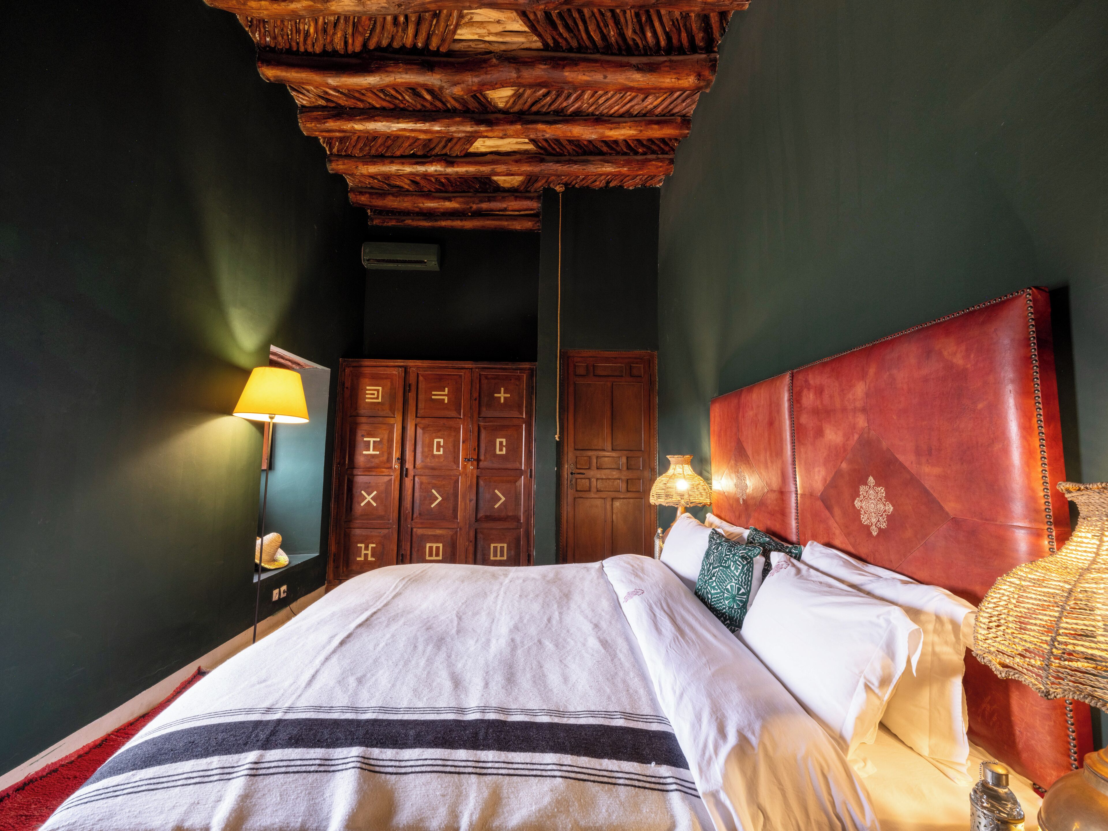 A bedroom with a wooden ceiling, a bed with white linens, a red headboard, and a wall-mounted lamp.