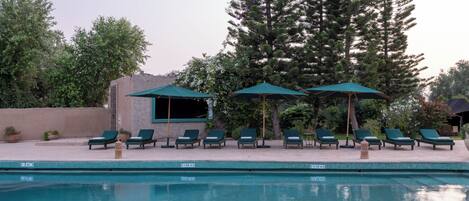 Zona de piscina con sillones de descanso y sombrillas, rodeada de árboles y un edificio.