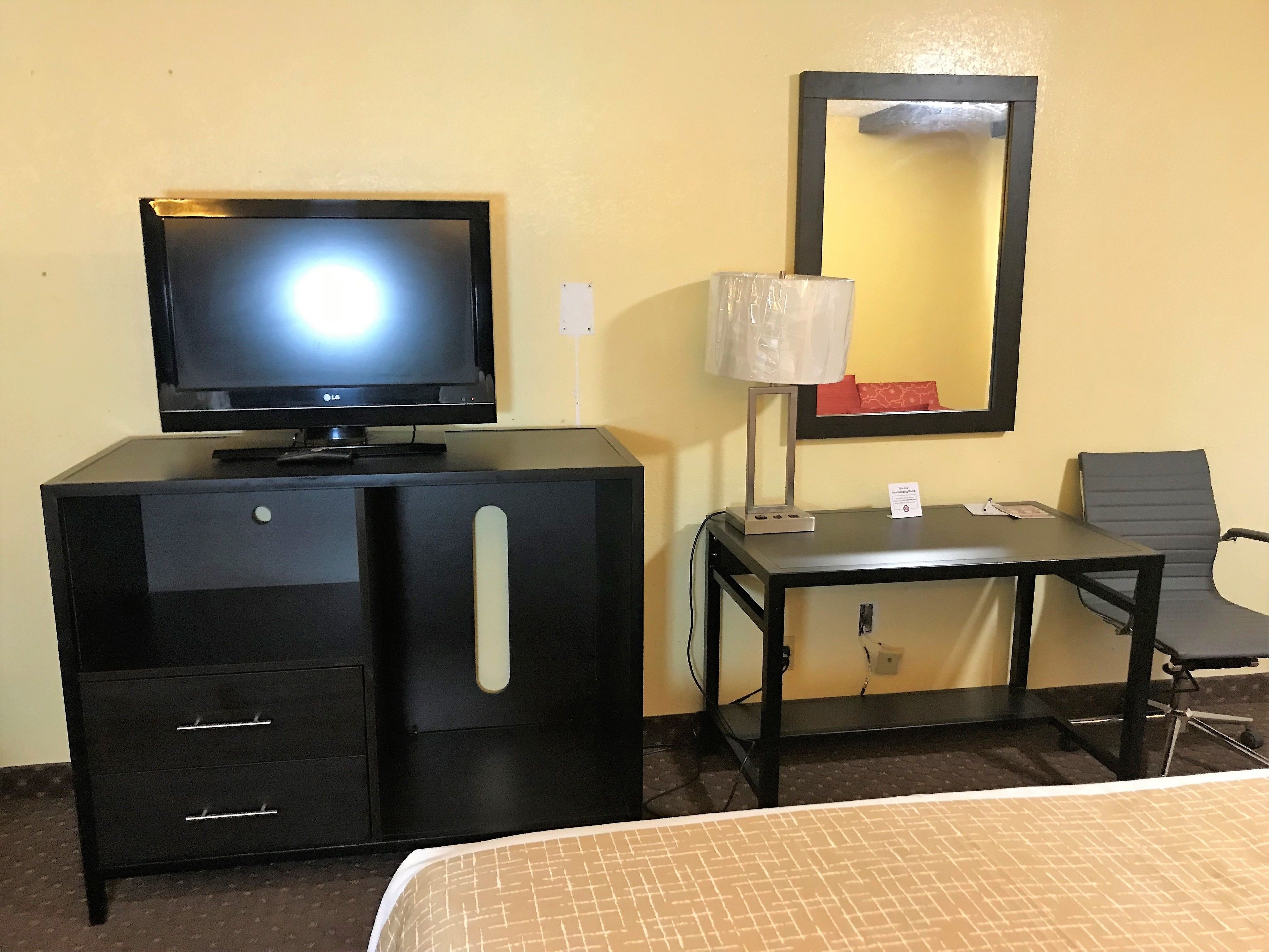 Ein Hotelzimmer mit Fernseher auf einer schwarzen Kommode, einem Schreibtisch mit Lampe, einem Sessel und einem Bett mit gemustertem Überwurf.