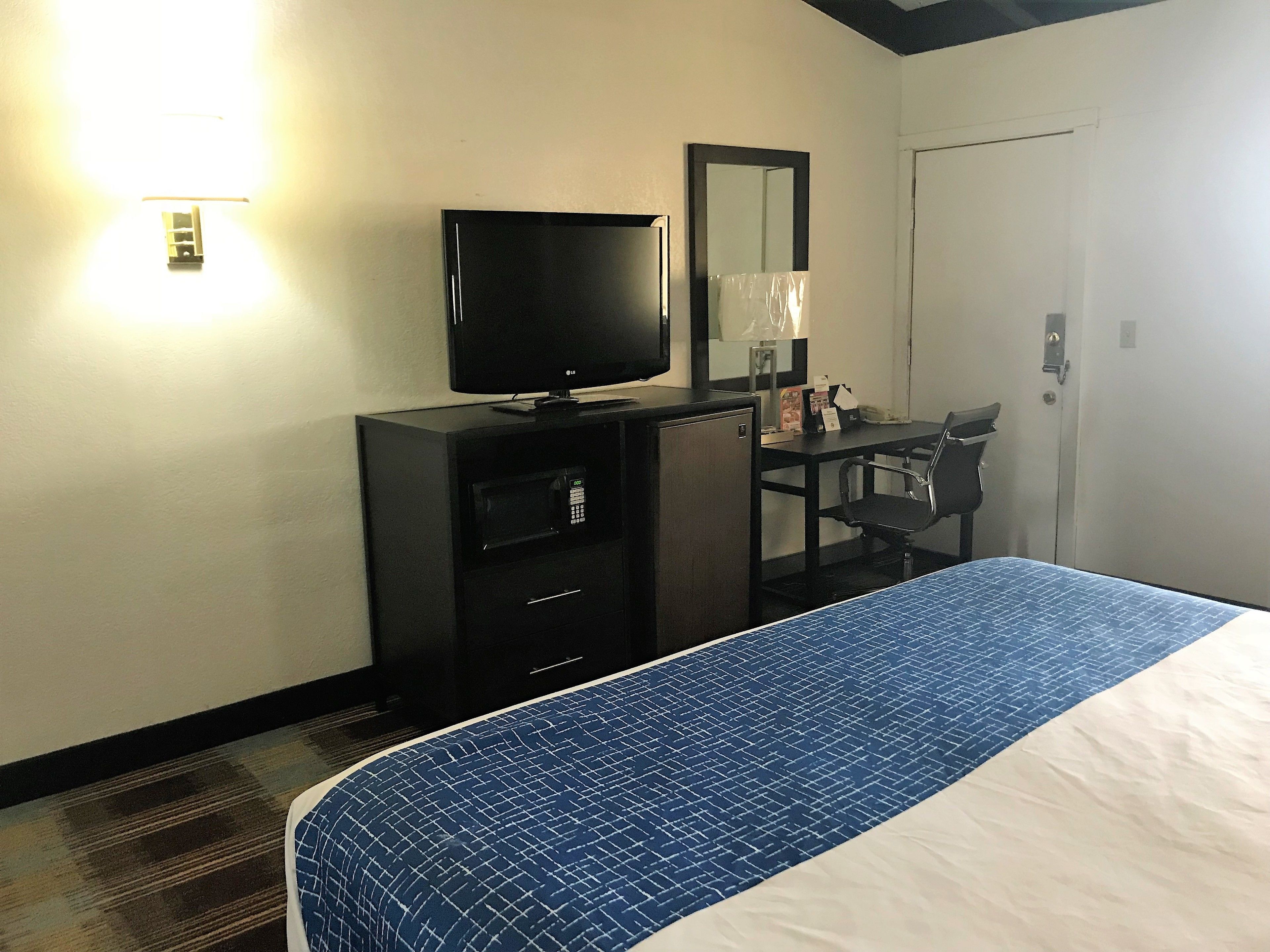 A hotel room with a bed, a desk with a chair, a television, a mirror, and a door.