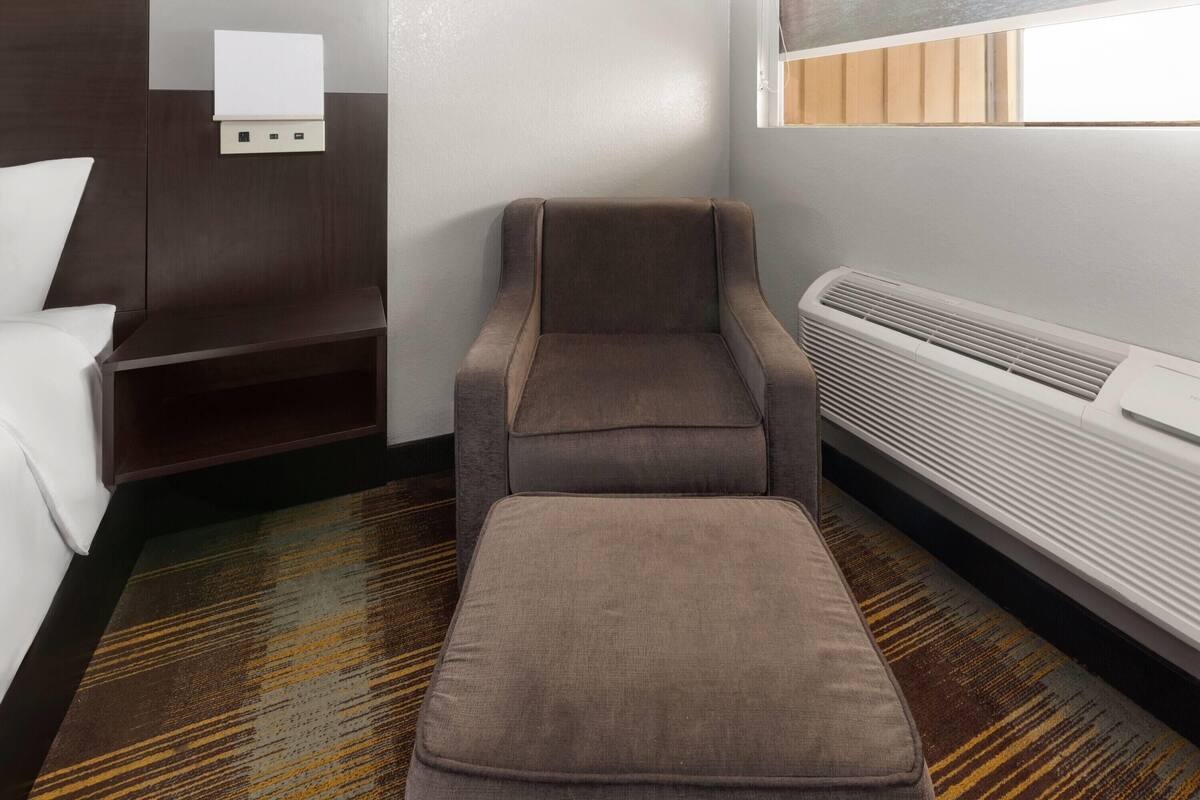 Une chambre d’hôtel avec un canapé marron, un pouf assorti, une petite table, une prise électrique fixée au mur et une fenêtre avec des stores.