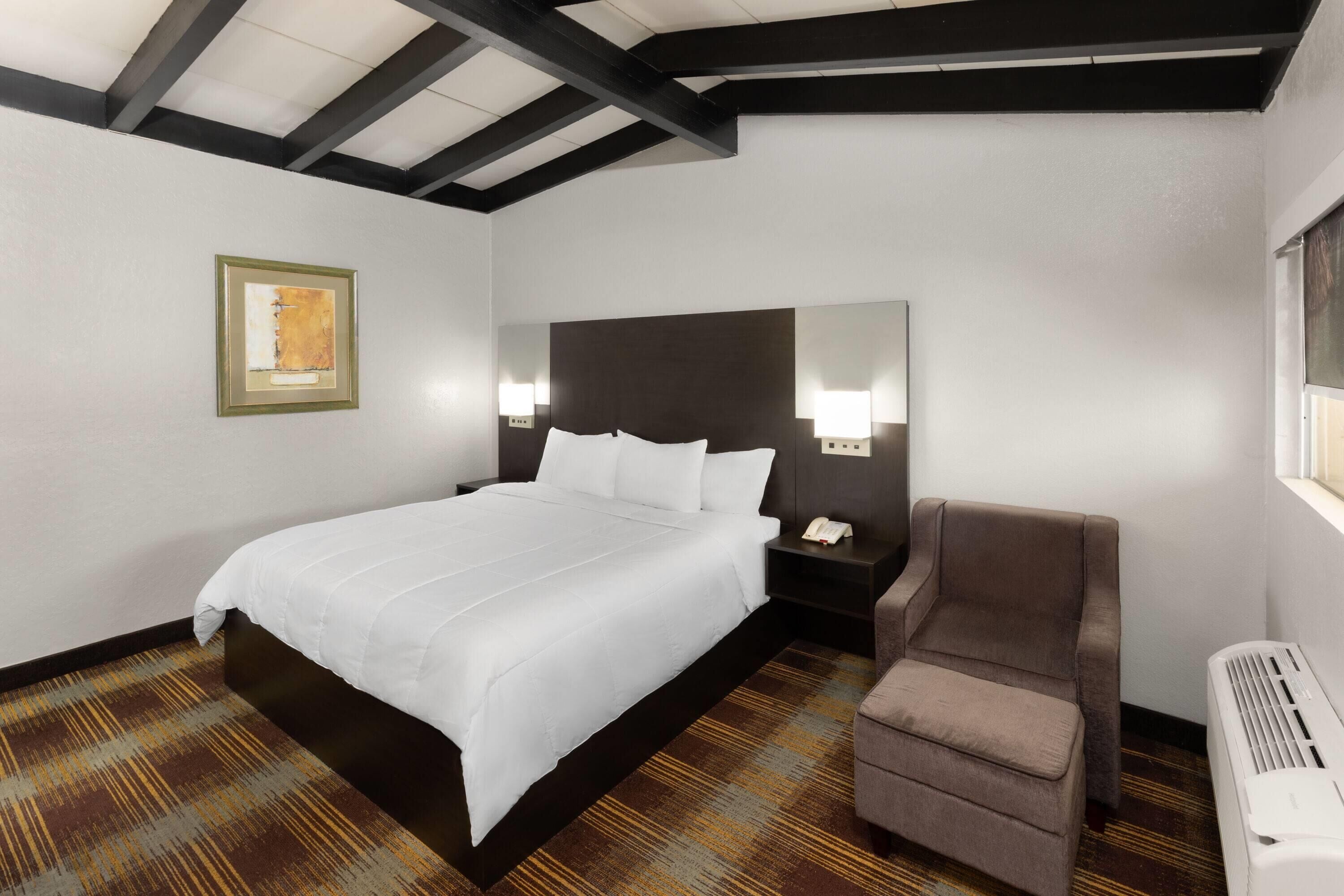 Una habitación de hotel con una cama grande, una silla, una mesita de noche con un teléfono, un espejo en la pared y un radiador.
