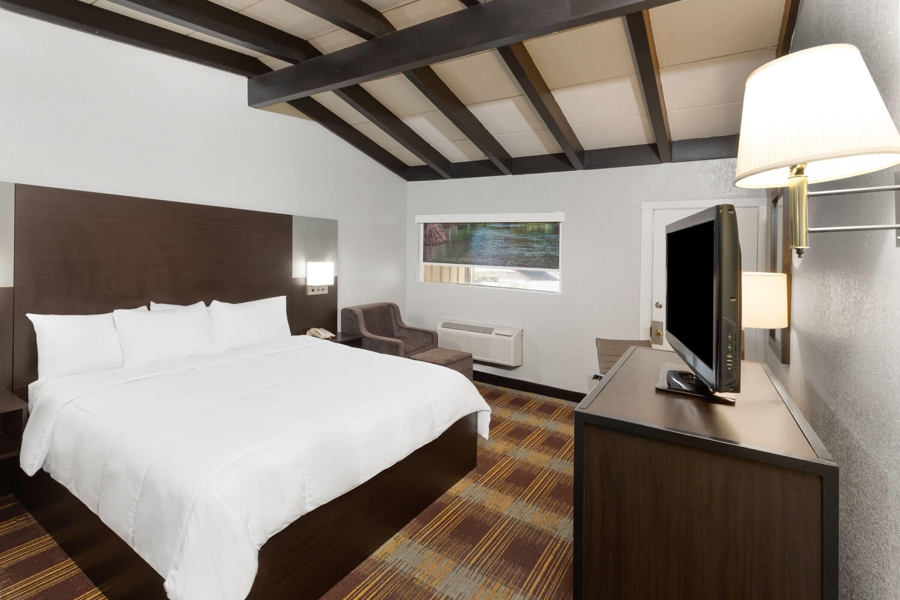 Quarto de hotel com uma cama grande, uma escrivaninha com televisão de tela plana, uma cadeira, uma janela com vista e teto com vigas aparentes.