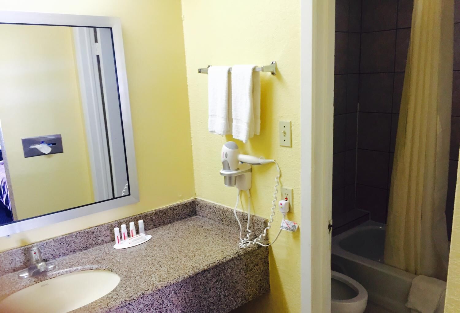 Un baño con encimera de granito, un lavabo, un espejo, un perchero con dos toallas, un secador de pelo y una cortina de ducha.