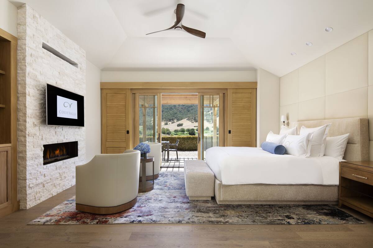Quarto com cama, televisão presa numa parede de tijolos, ventilador de teto e vista para o exterior.