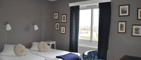 Habitación de hotel con una cama, dos lámparas de noche, una silla, una mesita y cuadros en la pared.