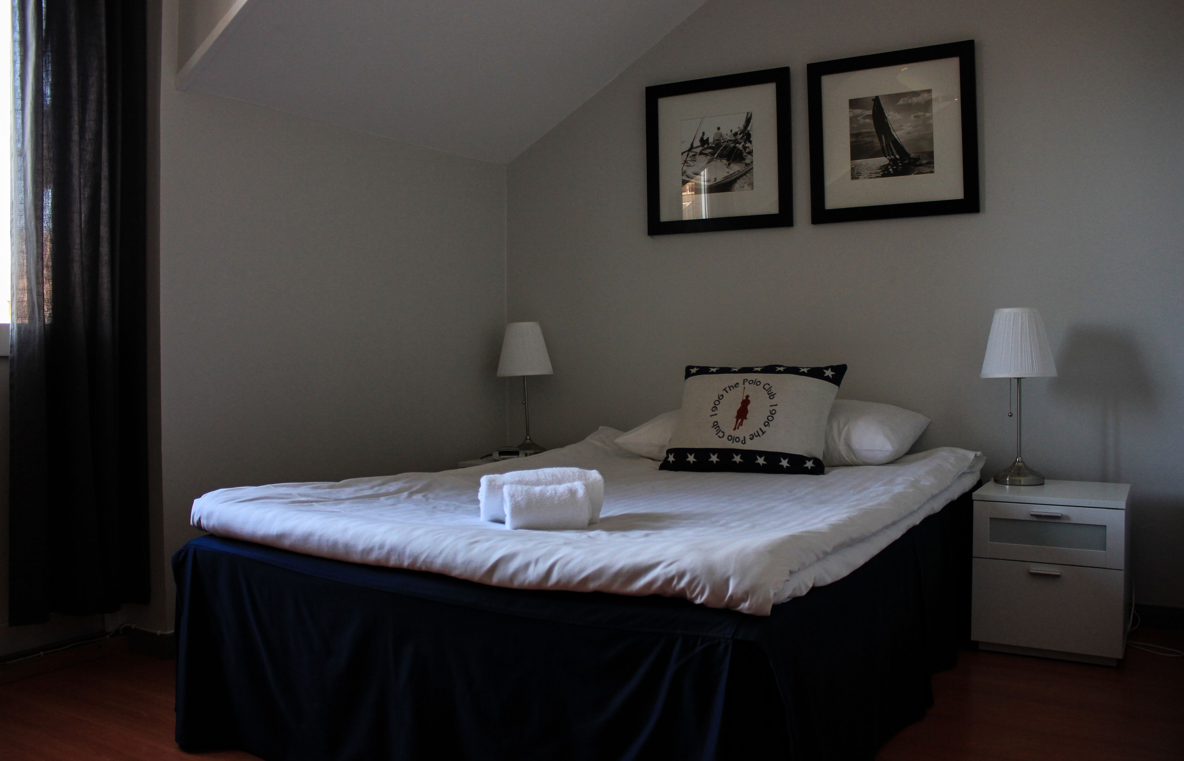 Un lit bien fait, recouvert d’une couvre-lit bleu foncé, avec deux lampes de chevet, une table de chevet et deux tableaux encadrés accrochés au mur.
