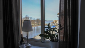 A table with a bottle of wine, two glasses, a lamp, and a potted plant by a window overlooking a river.