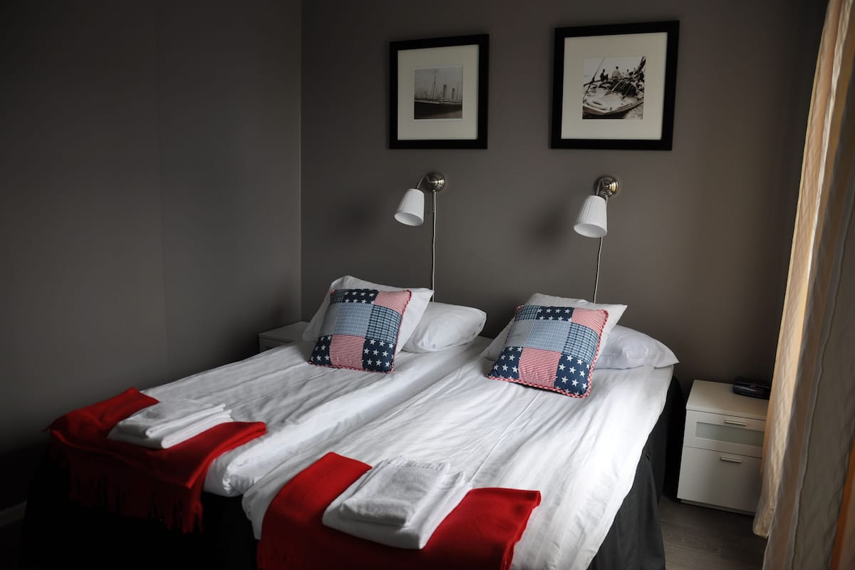 Un lit double avec du linge de lit blanc et des touches de rouge, deux lampes de chevet, une table de chevet avec une radio et deux tableaux encadrés au mur.