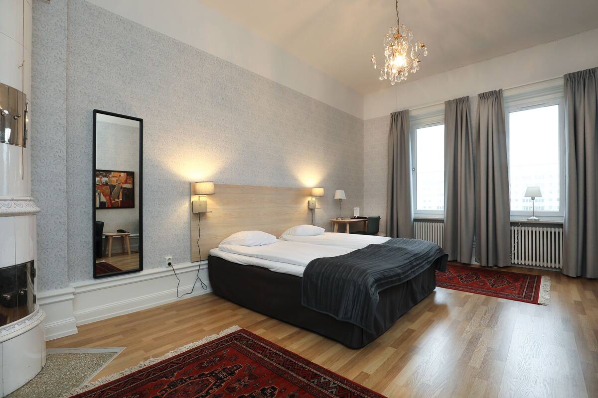 Ein Schlafzimmer mit einem Bett, einem großen Spiegel, Nachttischlampen, einer Kristallleuchter und Blick auf die Stadt durch große Fenster.