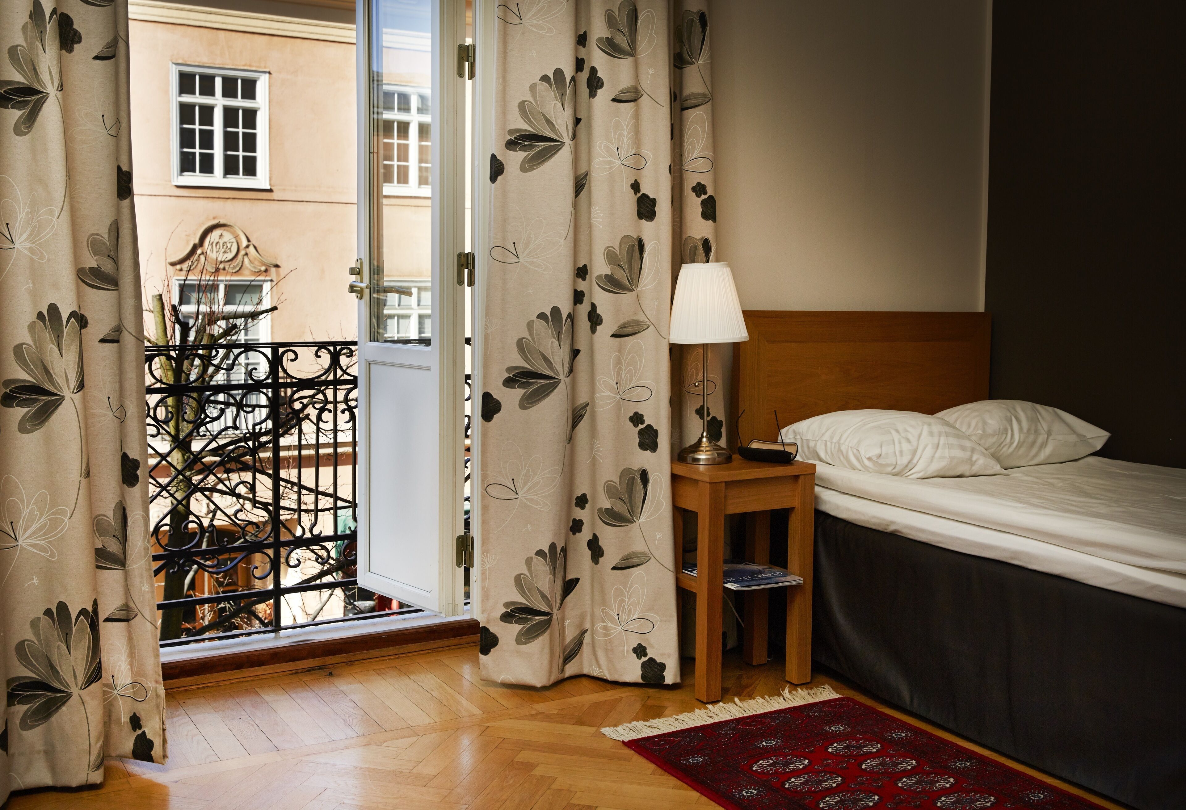 A bedroom with a bed, a nightstand with a lamp, a red rug, and a view of a balcony with a wrought iron gate.