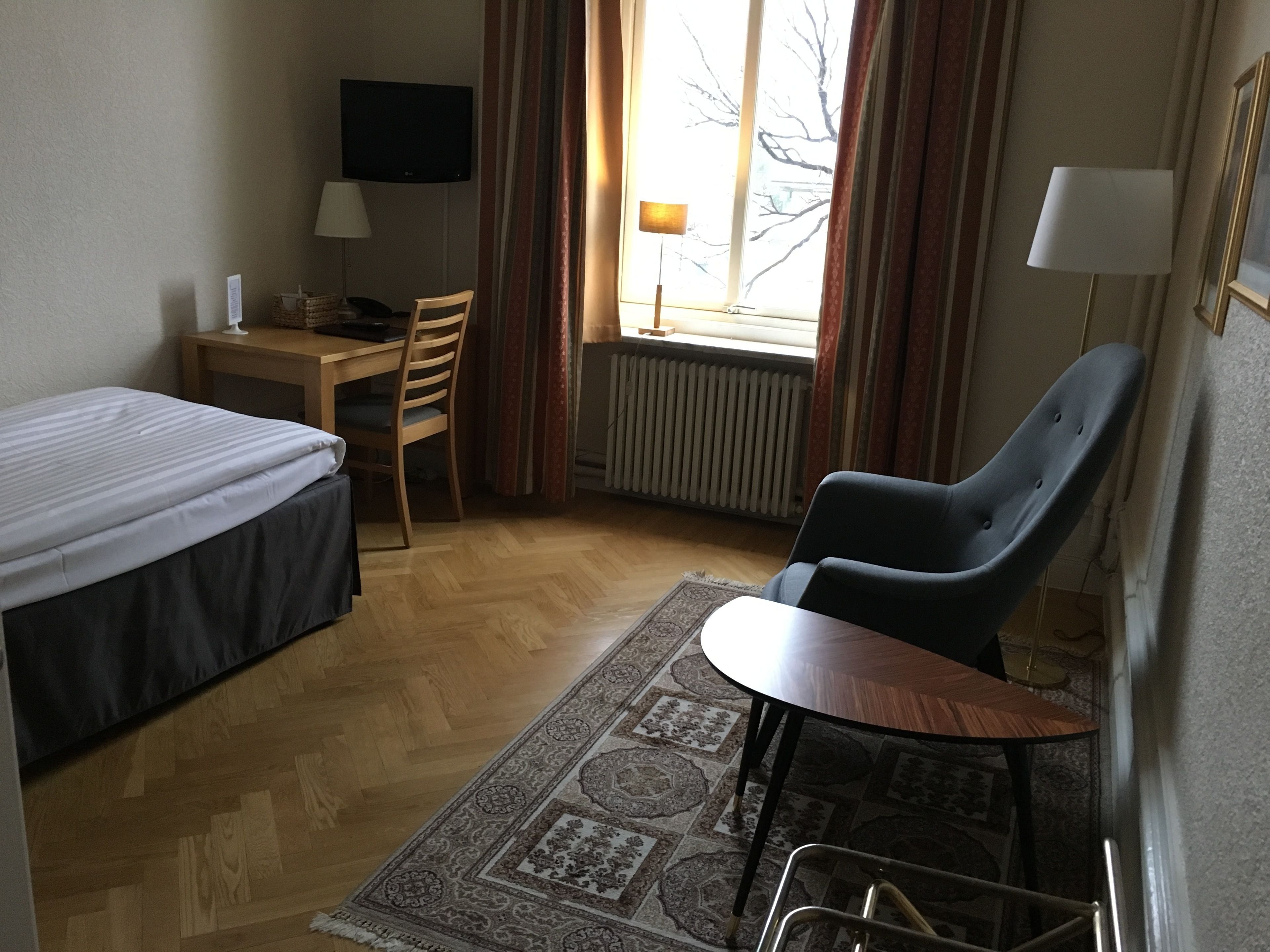 A hotel room with a bed, a desk, a chair, a small table, a TV, and a window with curtains.