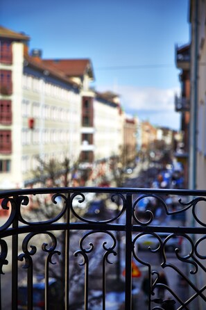 A balcony with wrought iron railings overlooking a street with buildings and parked cars.