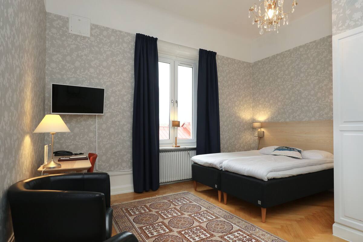 Ein Schlafzimmer mit einem Bett, einem Schreibtisch mit Lampe, einem Stuhl, einem Fernseher und einem Fenster mit Vorhängen.