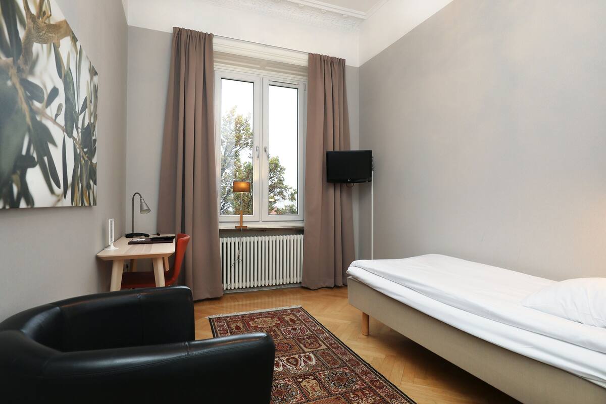 Ein Schlafzimmer mit Bett, Schreibtisch, Stuhl, Fernseher und einem Fenster mit Vorhängen.