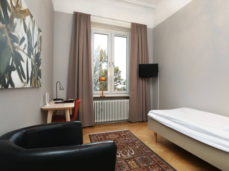 A bedroom with a bed, a desk, a chair, a TV, and a window with curtains.