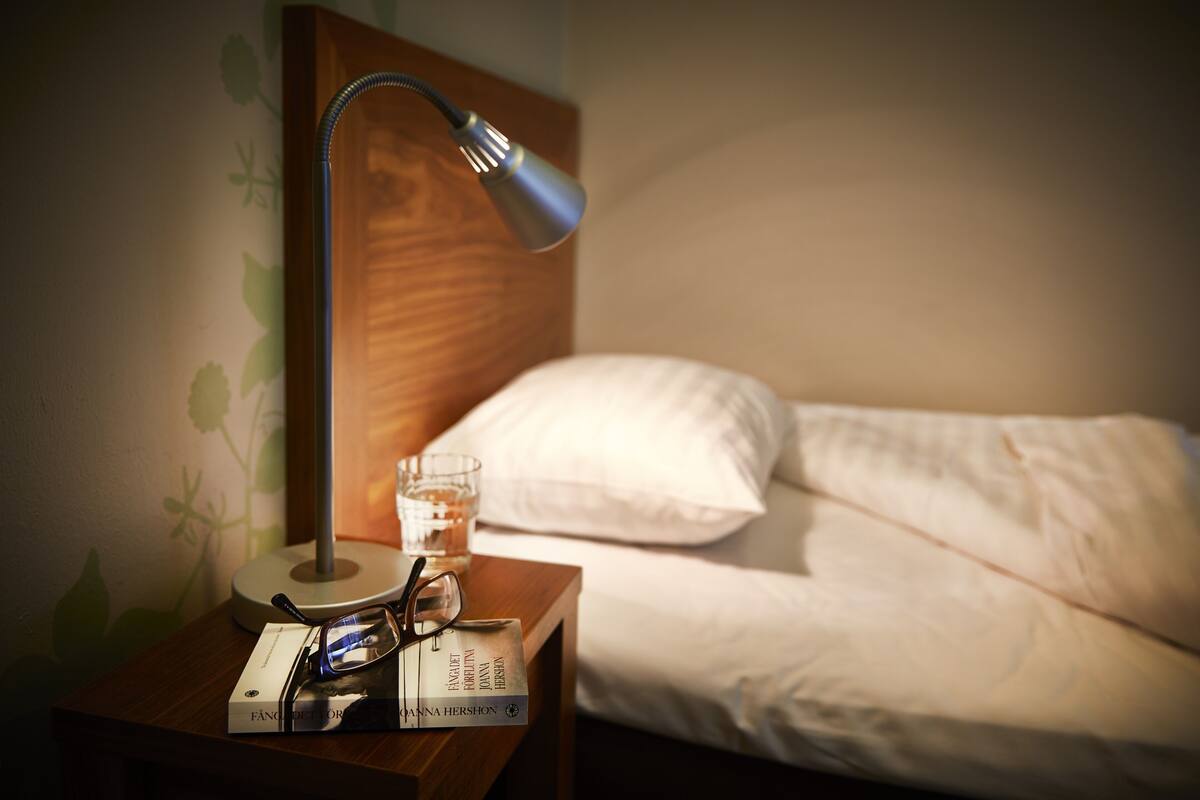 Ein ordentlich bezogenes Bett mit weißen Leinen, ein Nachttisch mit Lampe, einem Glas Wasser und einem Buch.