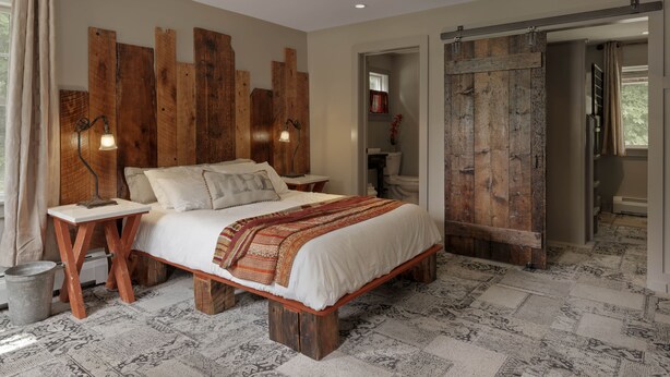 A bedroom with a wooden bed, a nightstand, a lamp, and a large wooden wall panel. - Mad River Barn (Waitsfield)