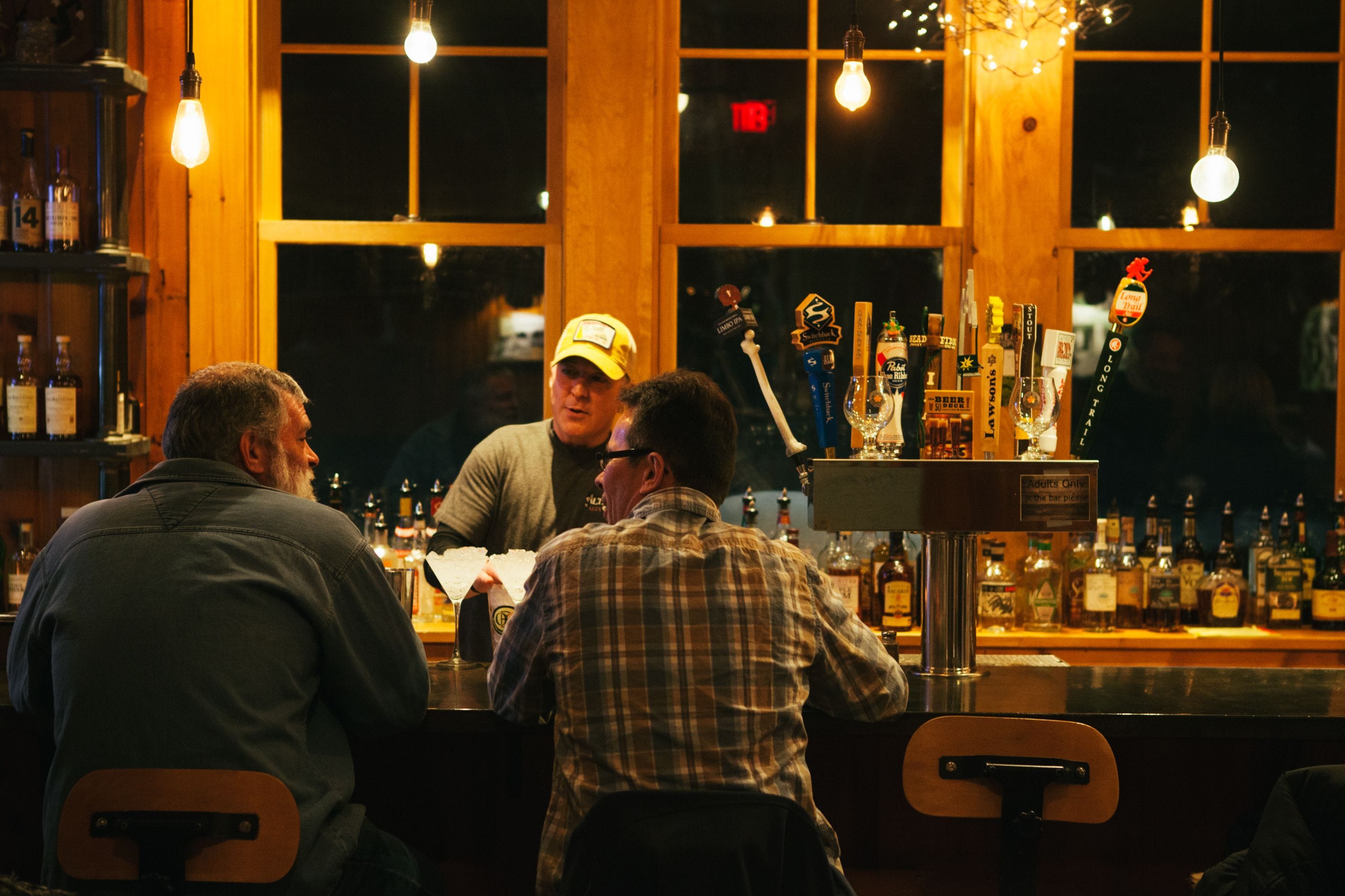 A bar with wooden walls and large windows, featuring a variety of bottles and a tap.