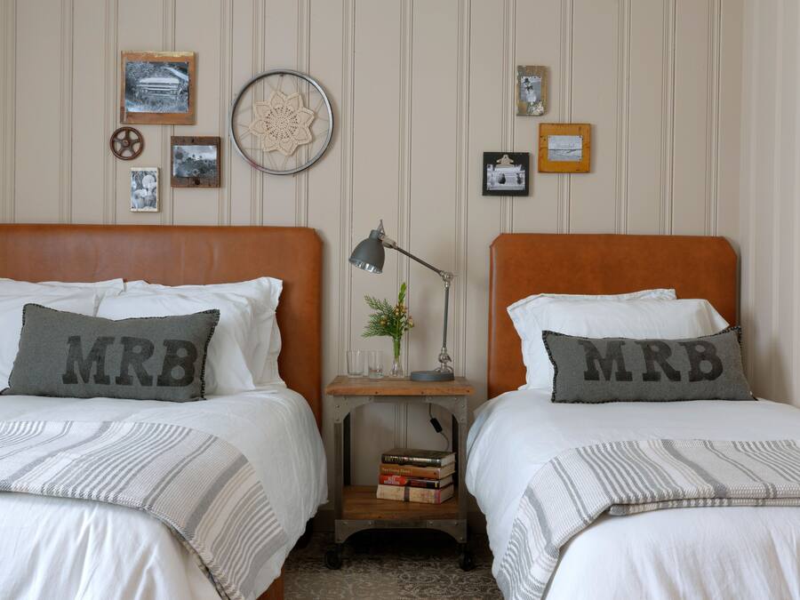 Un dormitorio con dos camas, cada una con una almohada etiquetada “MRB”. Hay cuadros en la pared, una mesita con una lámpara y una estantería con libros.