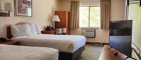 Quarto de hotel com duas camas, uma escrivaninha com televisão, uma luminária e um quadro na parede.