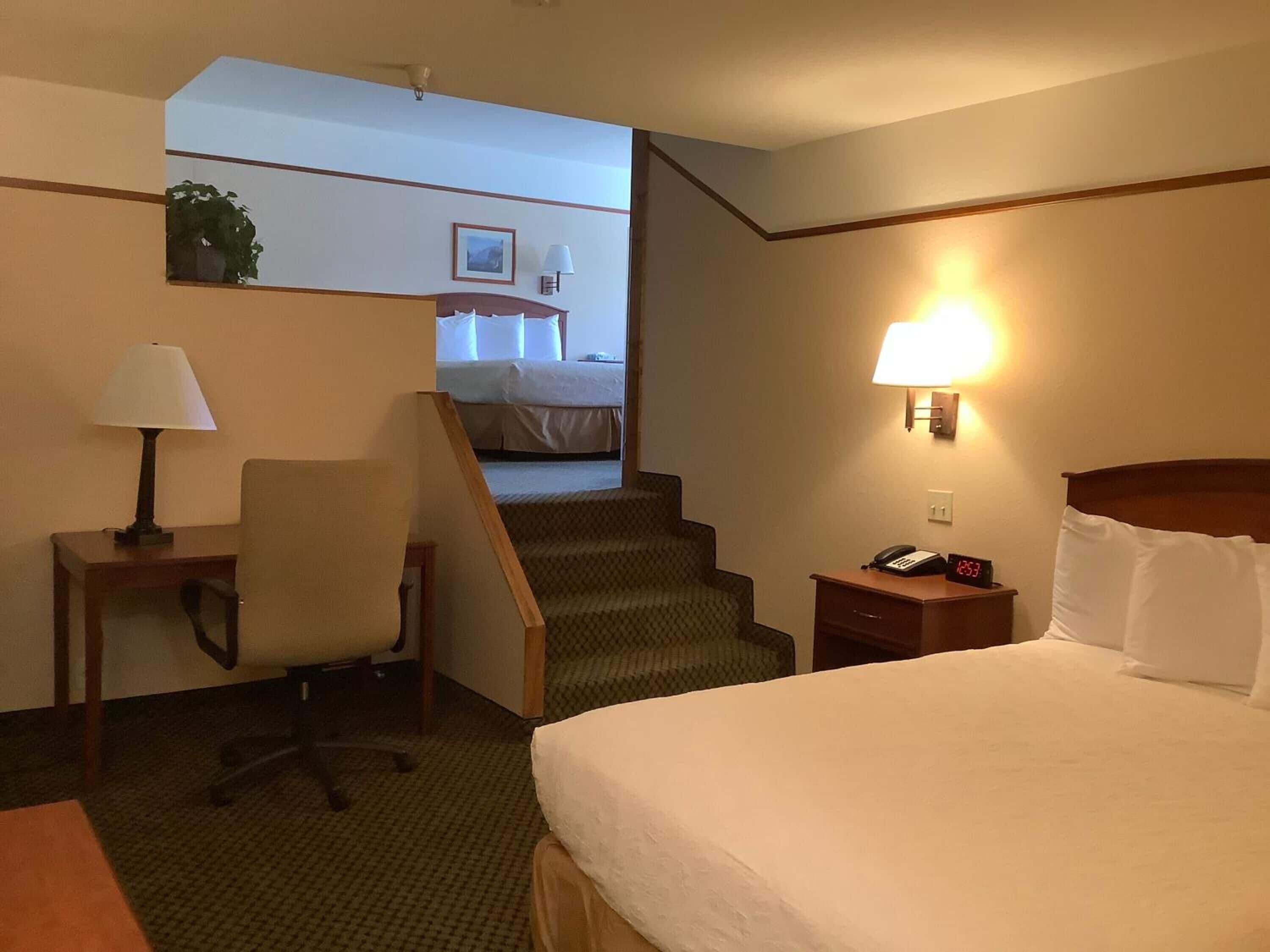 A hotel room with a bed, a desk with a lamp, a chair, a nightstand with a phone, and a staircase leading to another room.