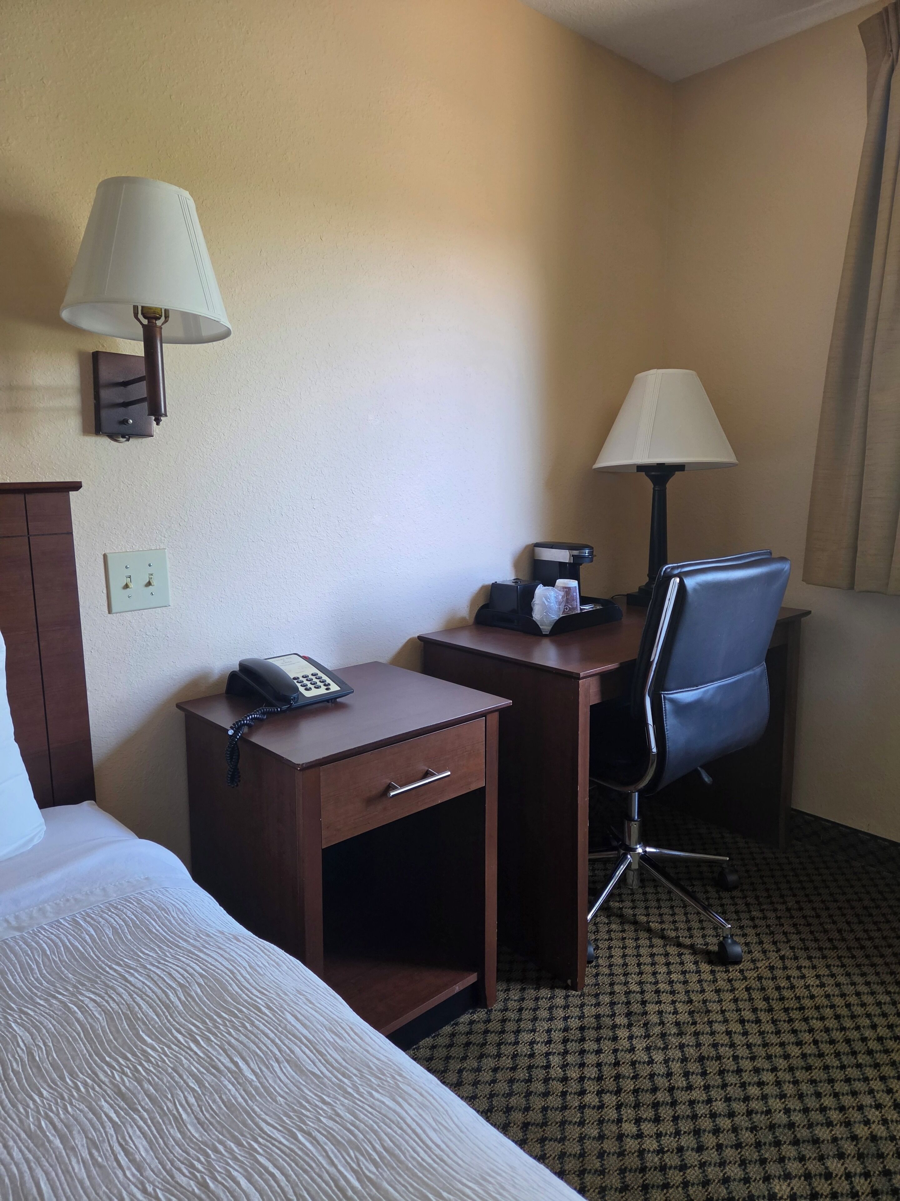 A hotel room with a bed, a desk with a telephone, a chair, and two wall-mounted lamps.