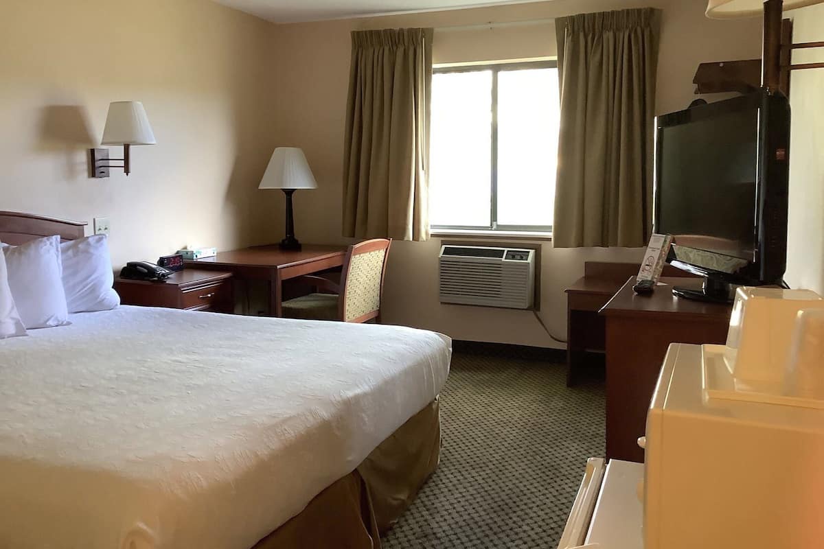 A hotel room with a bed, desk, chair, TV, air conditioning unit, and curtains.
