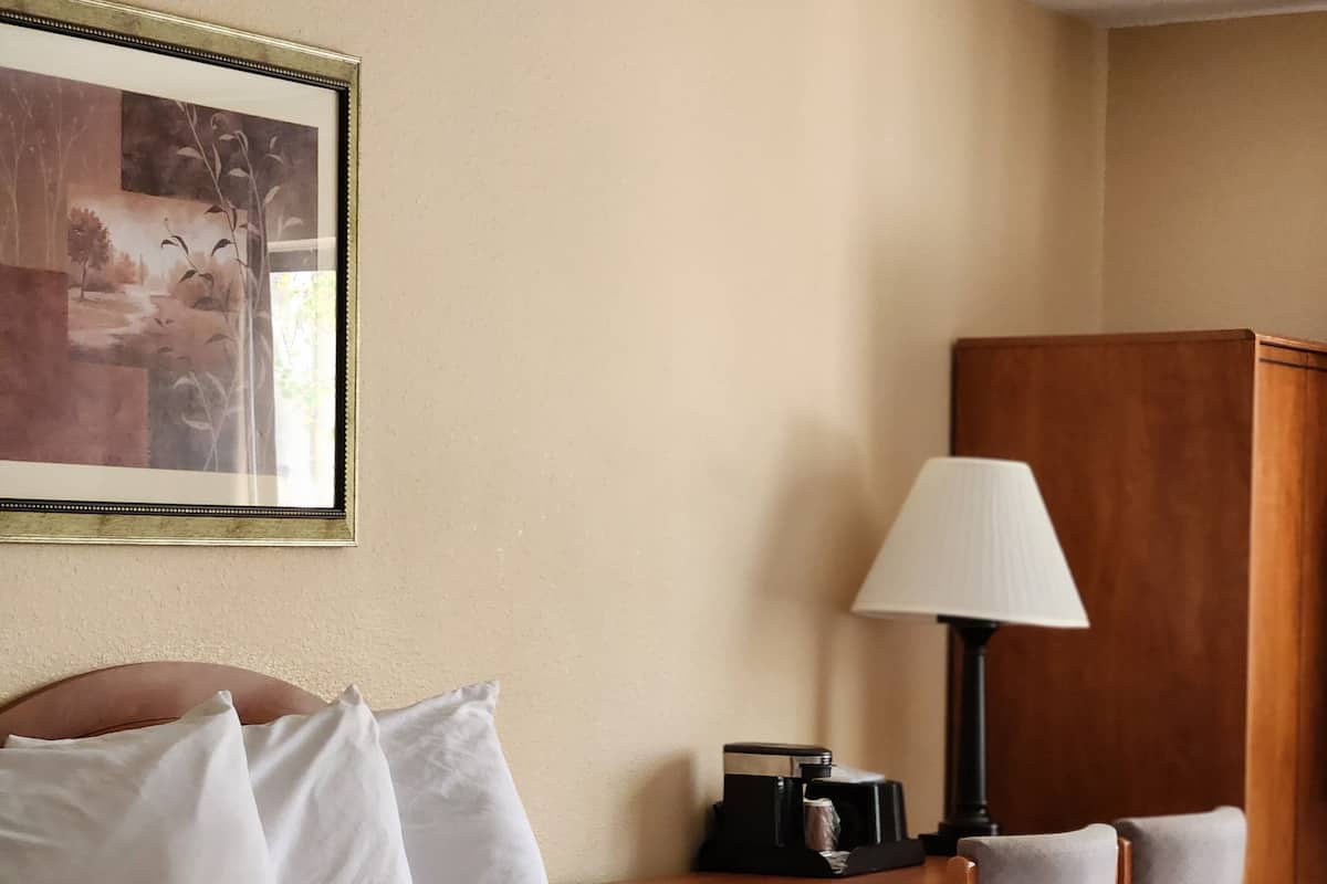 A hotel room with a bed, pillows, a nightstand with a lamp, a wooden wardrobe, and a framed picture on the wall.