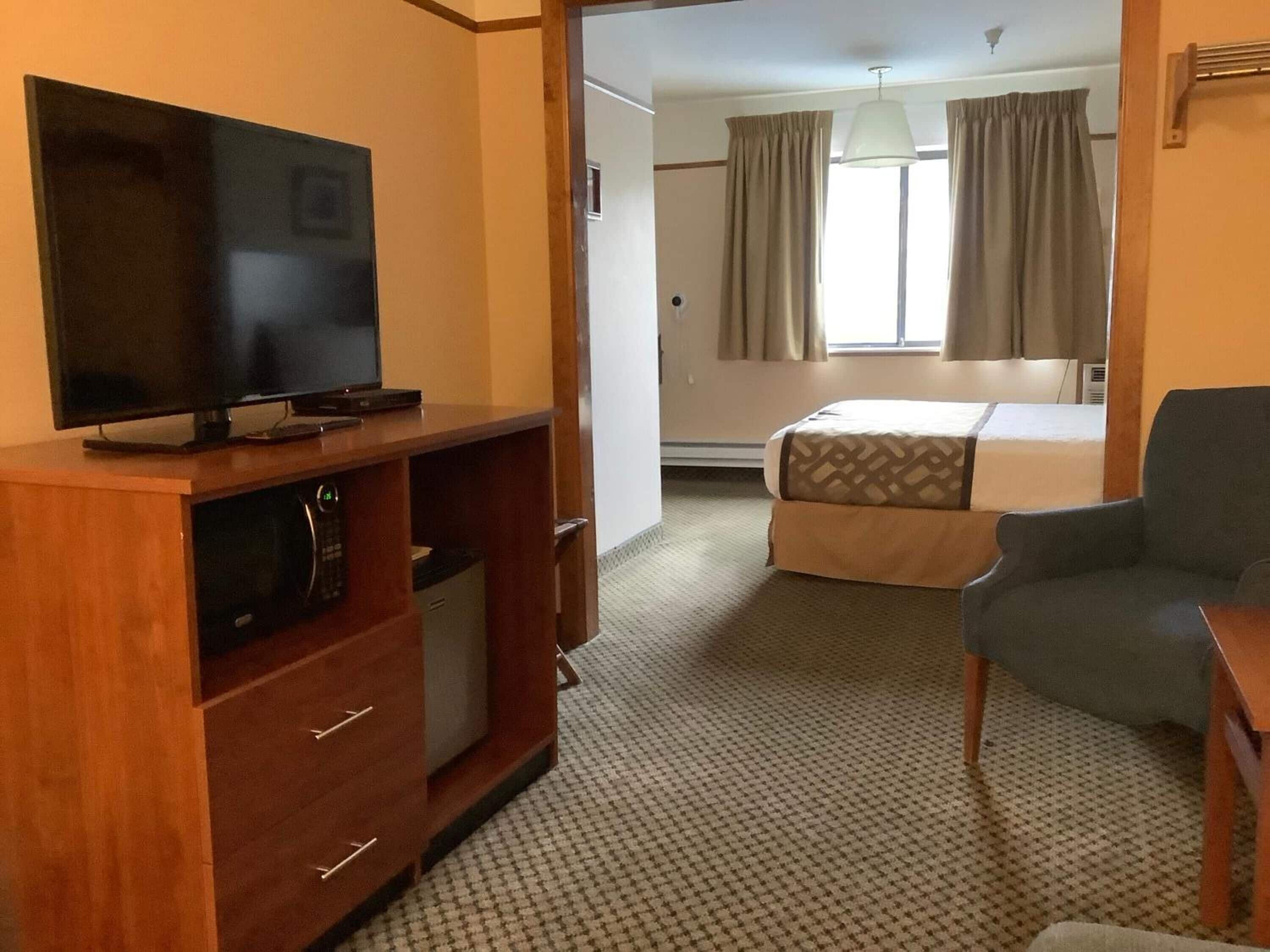 A hotel room with a television on a wooden stand, a bed with patterned bedding, a grey armchair, and a window with curtains.