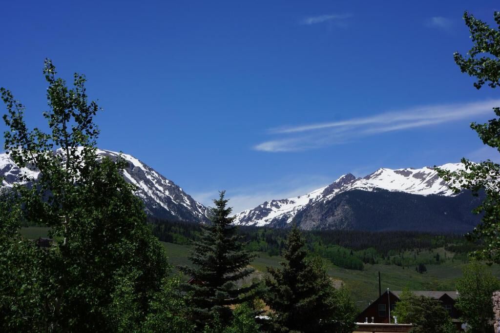A mountain landscape with snow-capped peaks, a clear sky, and a foreground of trees.
