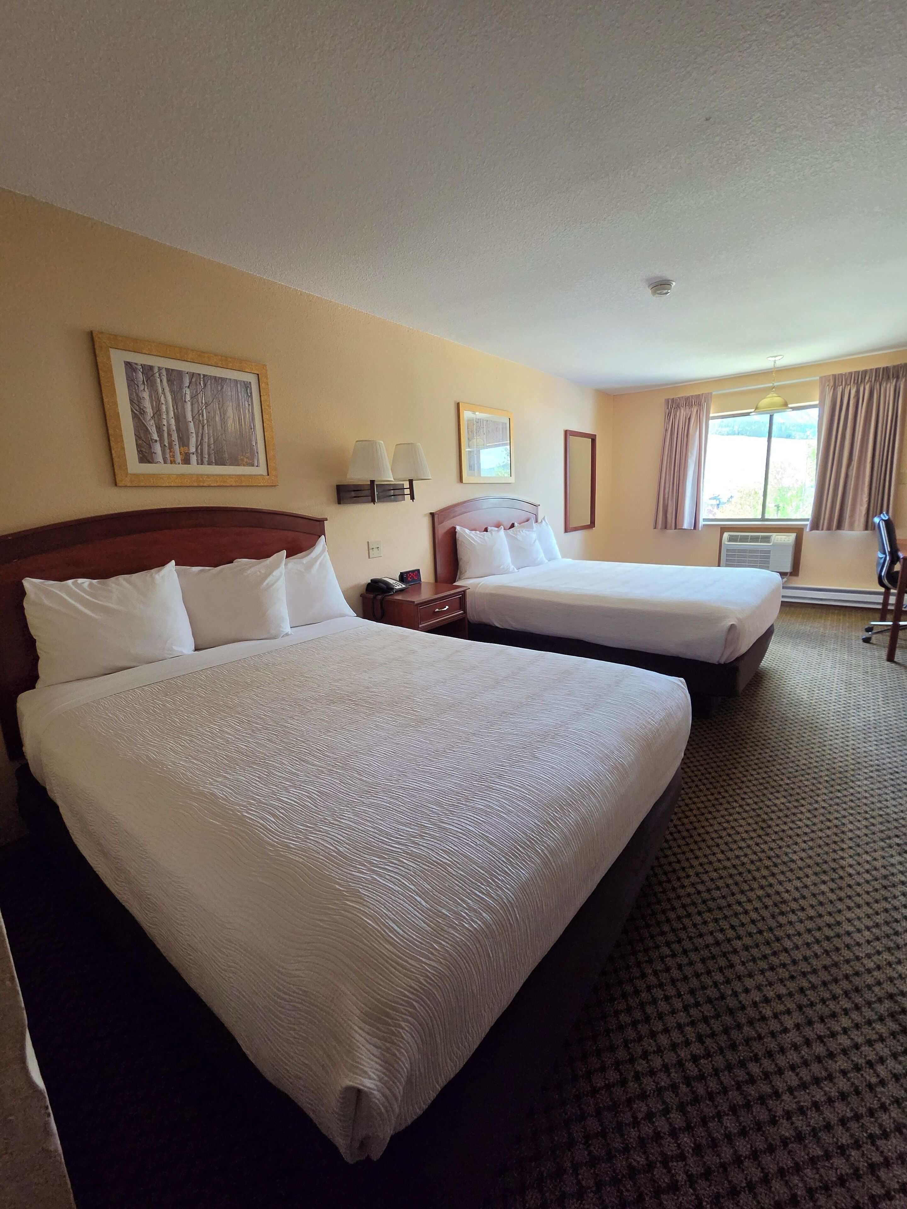 A hotel room with two beds, a bedside table with a lamp, a wall-mounted air conditioner, and a window with curtains.