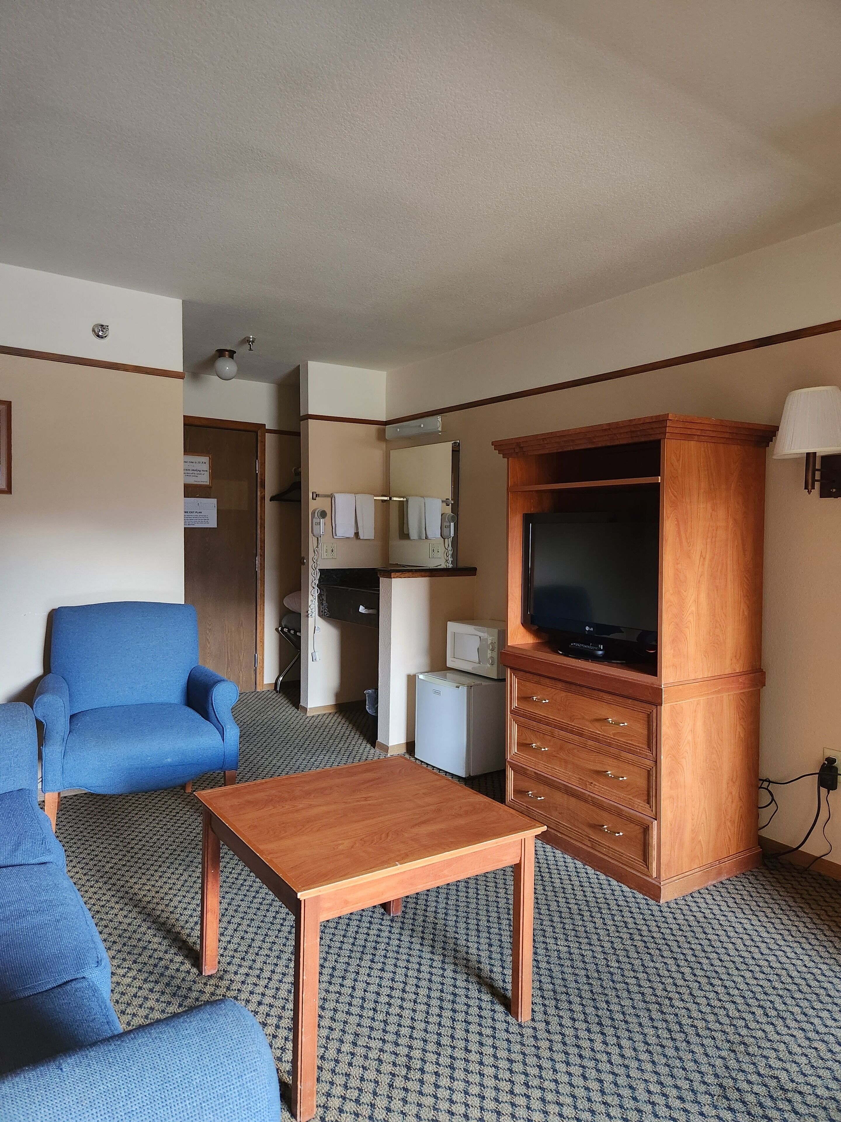 Une chambre d’hôtel avec un fauteuil bleu, une table basse en bois, un téléviseur posé sur un meuble en bois, un four à micro-ondes et un petit réfrigérateur.
