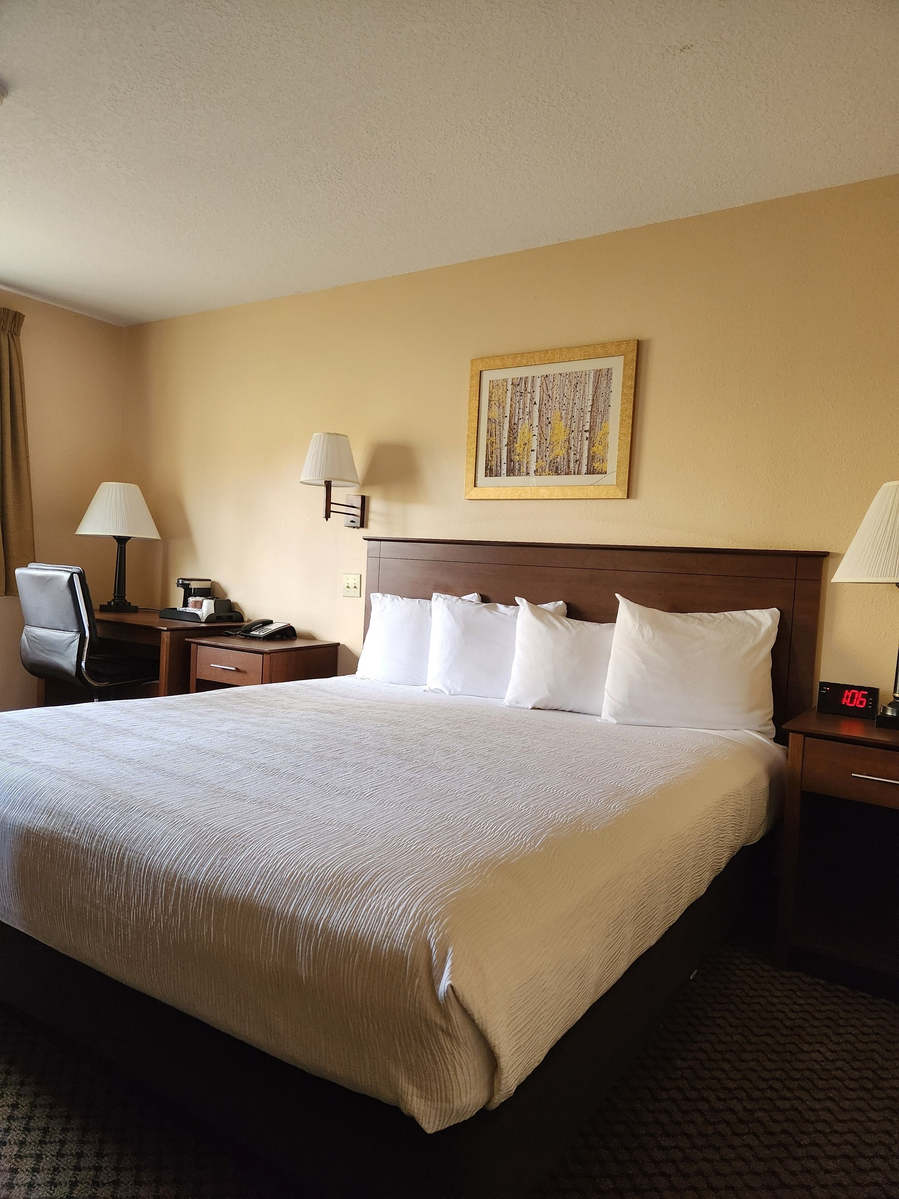 A hotel room with a large bed, two bedside tables, a desk with a chair, a lamp, a clock, and a framed picture on the wall.