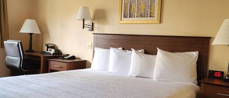 A hotel room with a large bed, two bedside tables, a desk with a chair, a lamp, a clock, and a framed picture on the wall.