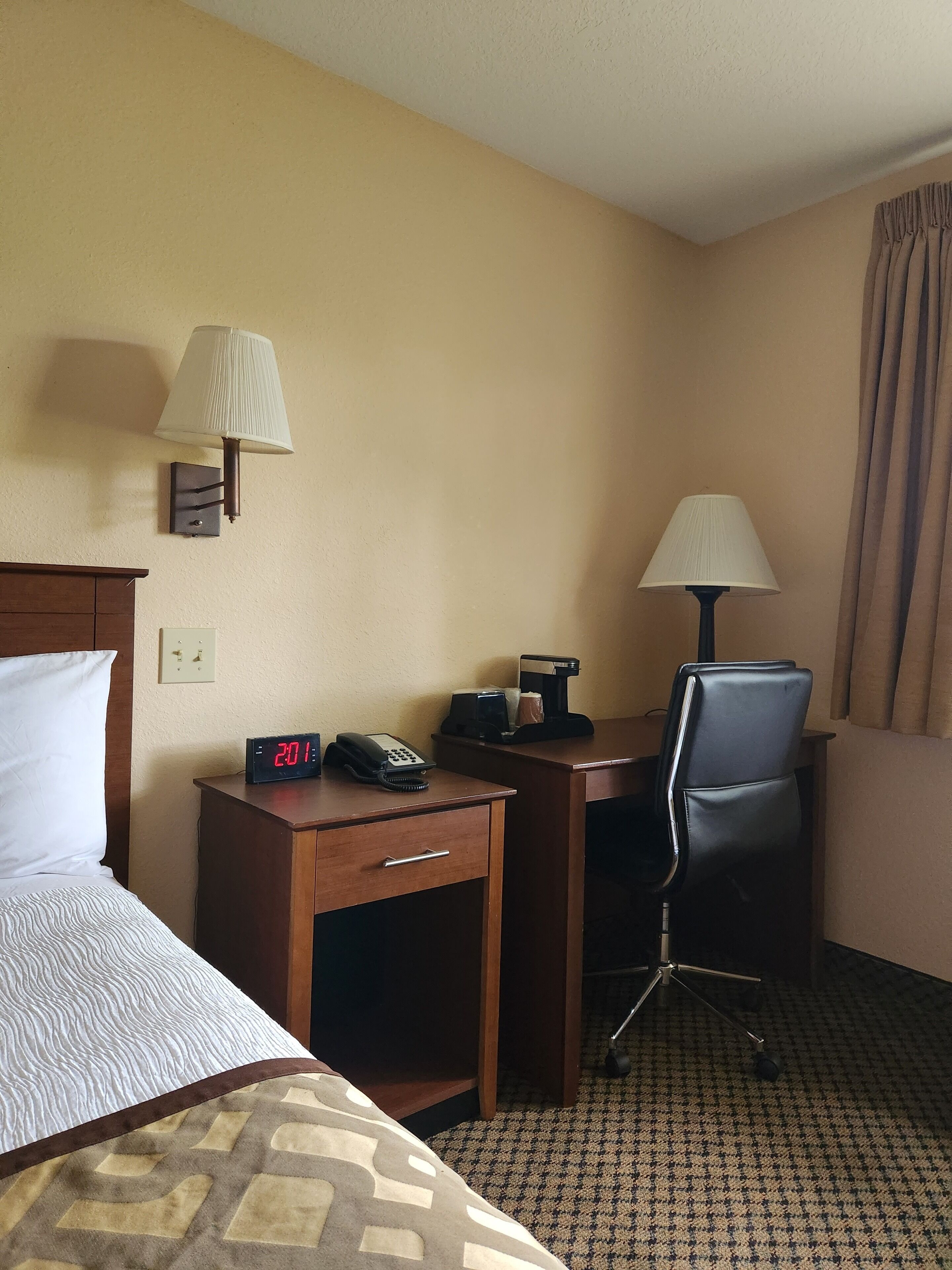 A hotel room with a bed, a desk with a lamp, a chair, a clock, and a coffee maker.