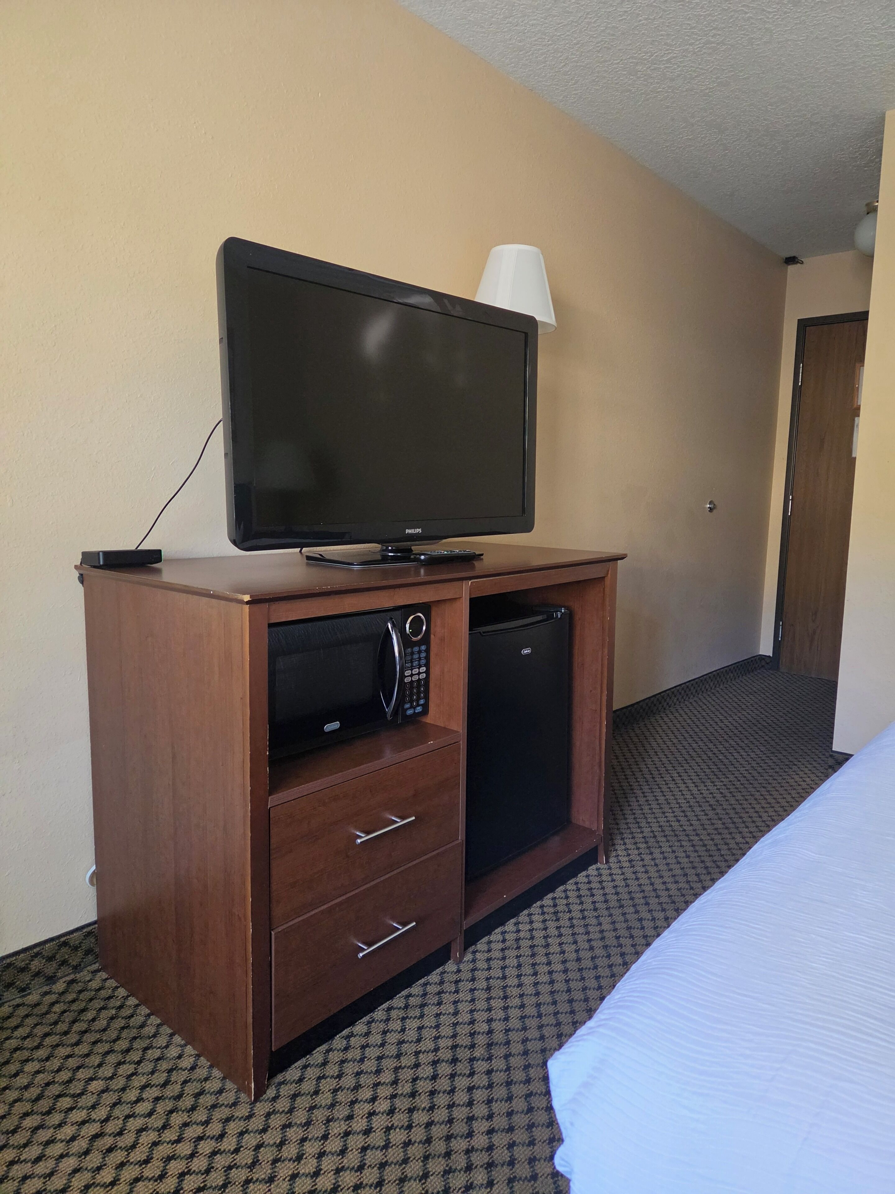Una habitación de hotel con televisión sobre un mueble de madera, microondas, refrigerador y una cama.