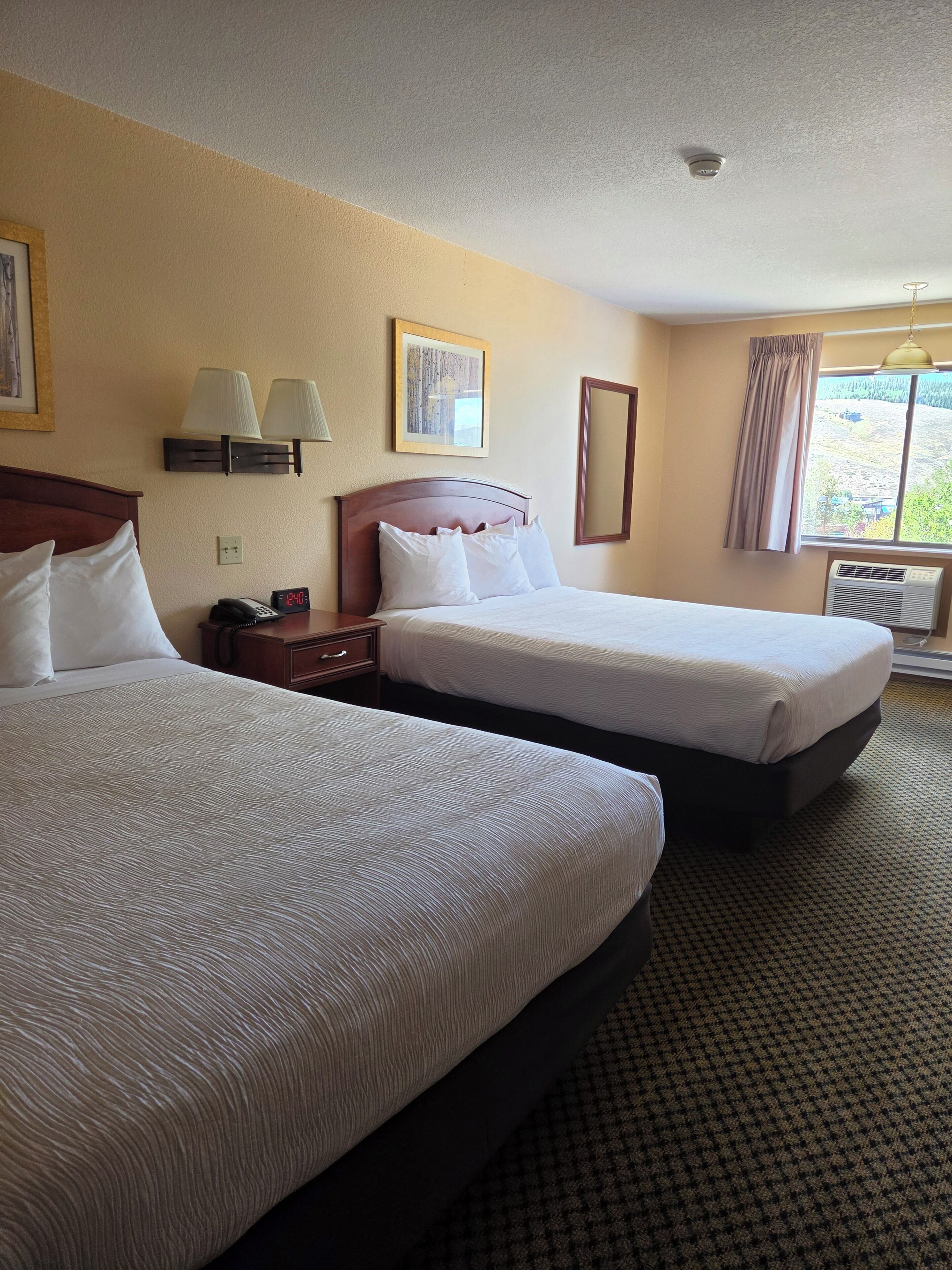 A hotel room with two beds, a bedside table with a lamp, a clock, and a window with curtains.