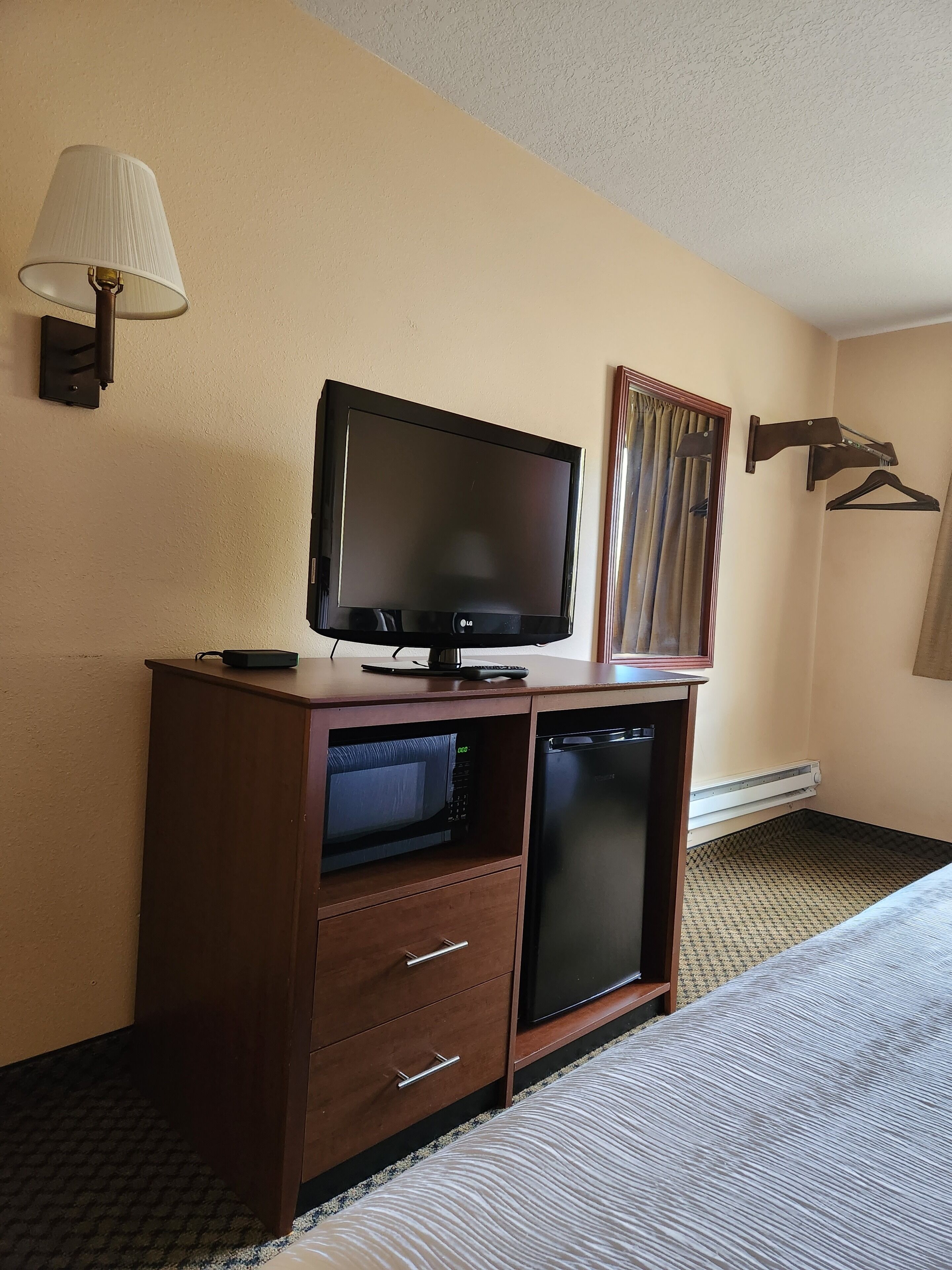 A hotel room with a bed, a TV on a wooden stand, a microwave, a small refrigerator, a lamp, and a coat rack.