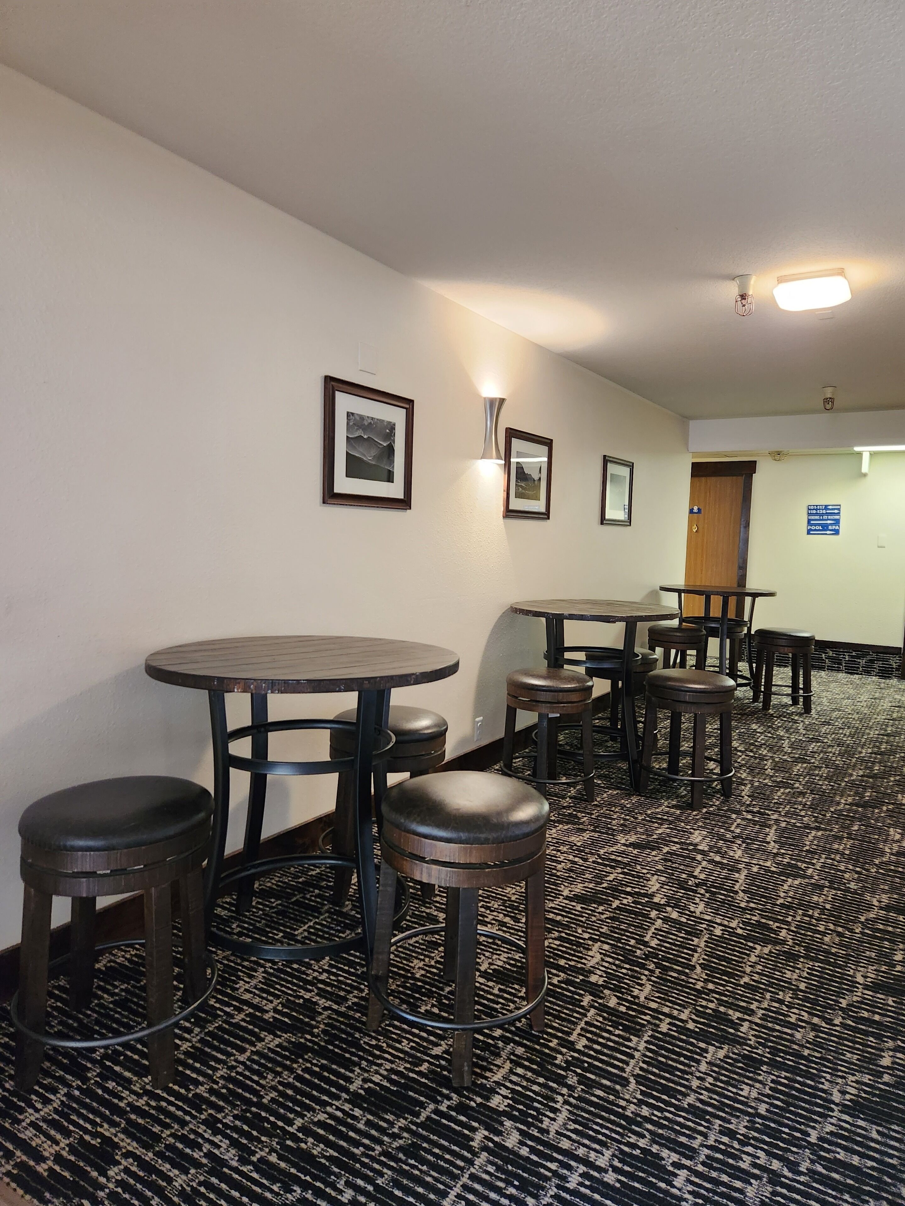 A hotel corridor with round tables and stools, framed pictures on the walls, and a carpeted floor.