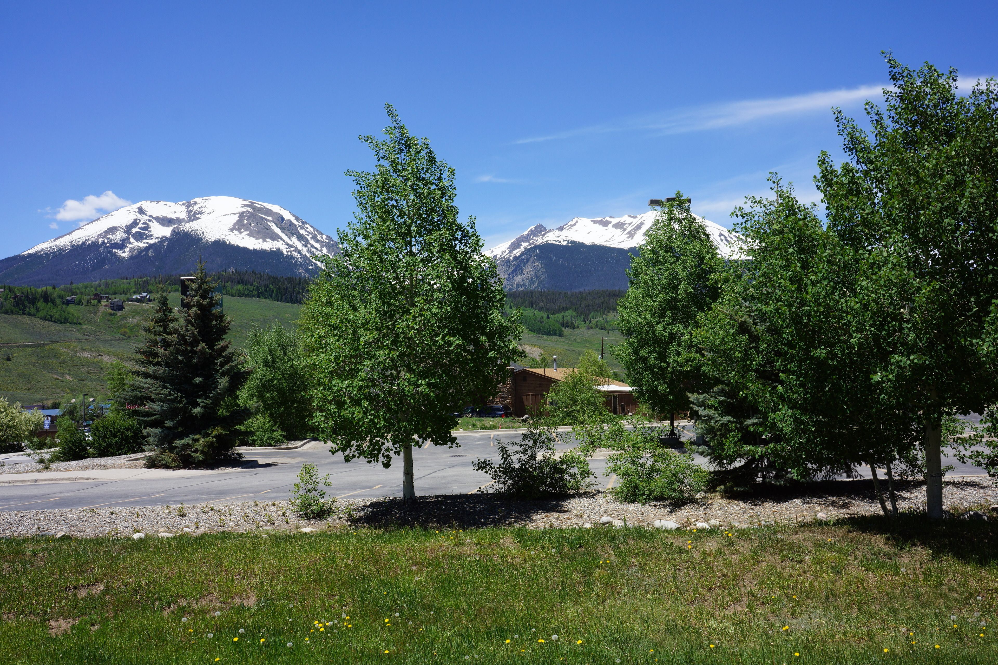 A mountain landscape with snow-capped peaks, a clear sky, and a parking area with trees and a building in the foreground.