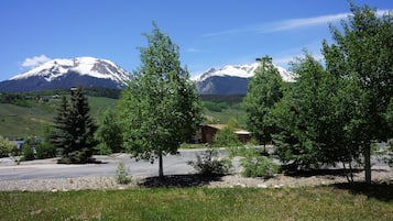 A mountain landscape with snow-capped peaks, a clear sky, and a parking area with trees and a building in the foreground.