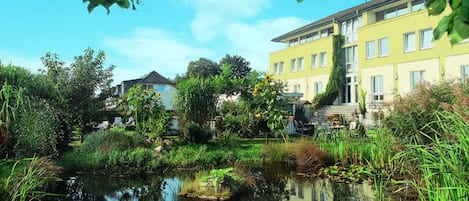 A serene lakeside view with a yellow building, surrounded by lush greenery and a calm water body reflecting the sky.