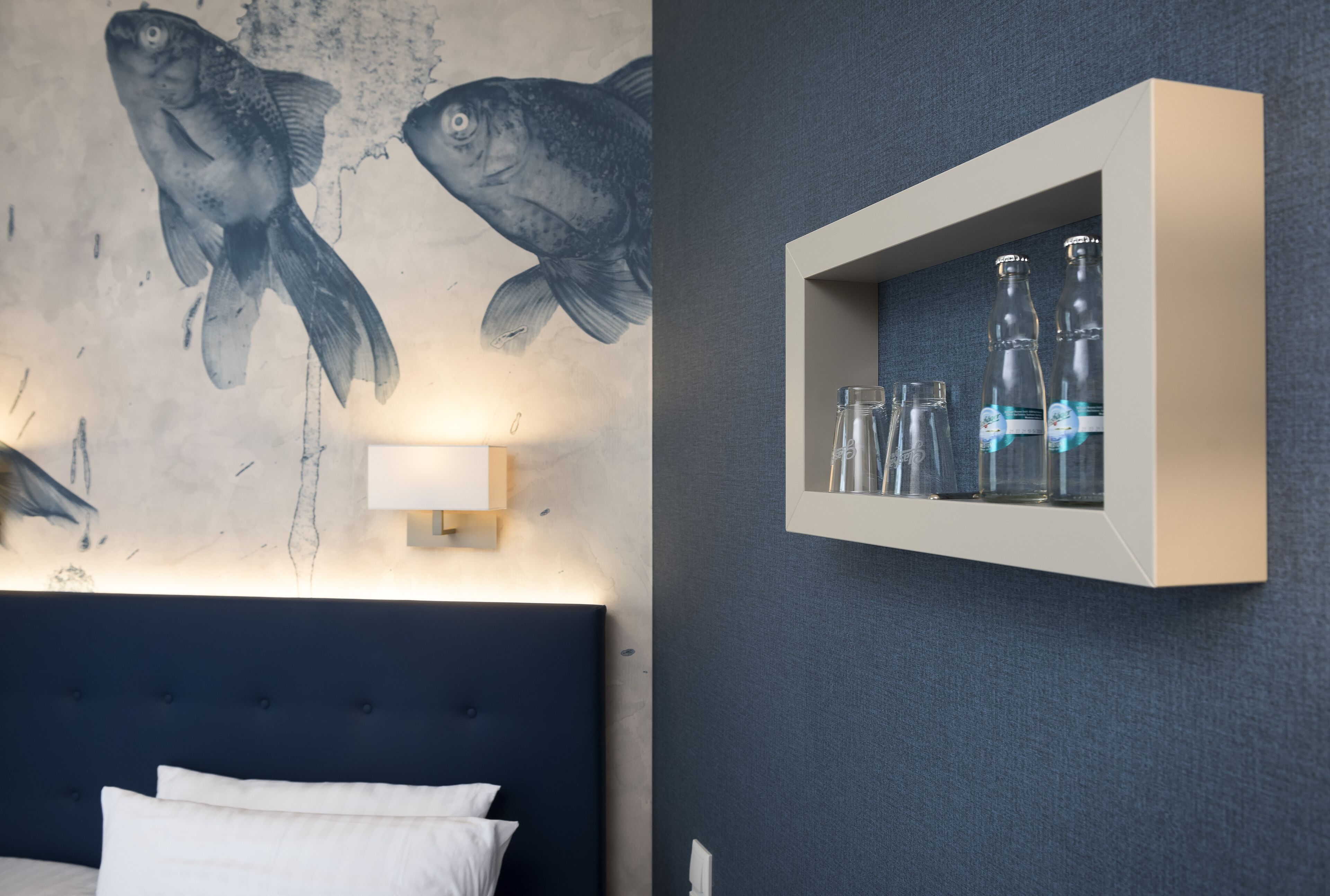 Une chambre avec une tête de lit bleu foncé, un lit recouvert de draps blancs et une étagère murale ornée de bouteilles et de verres.