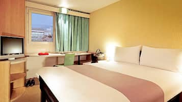 A hotel room with a bed, a desk with a television, a window with curtains, and a view of buildings outside.