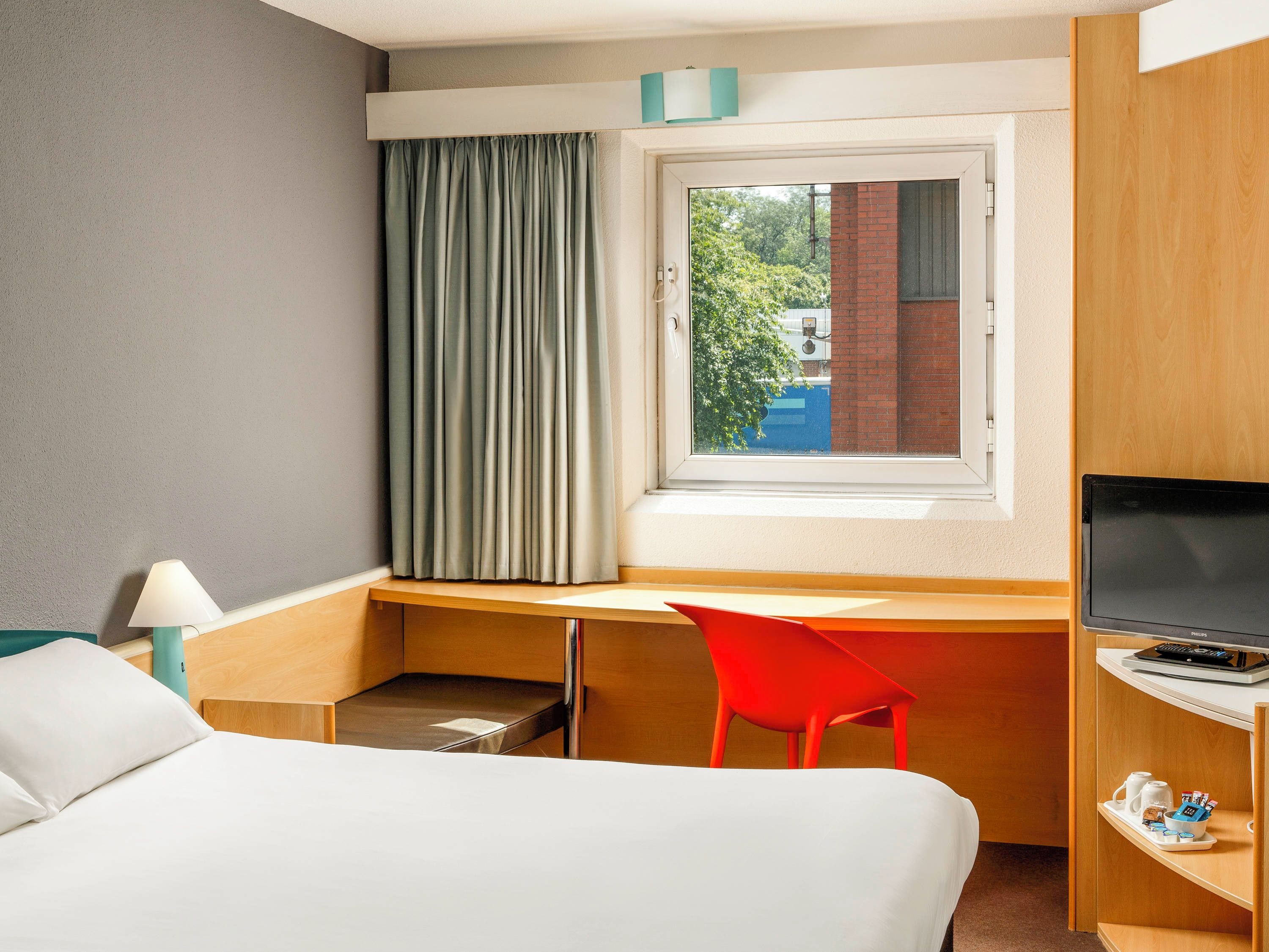 A hotel room with a bed, a desk, a red chair, a television, and a window with curtains.